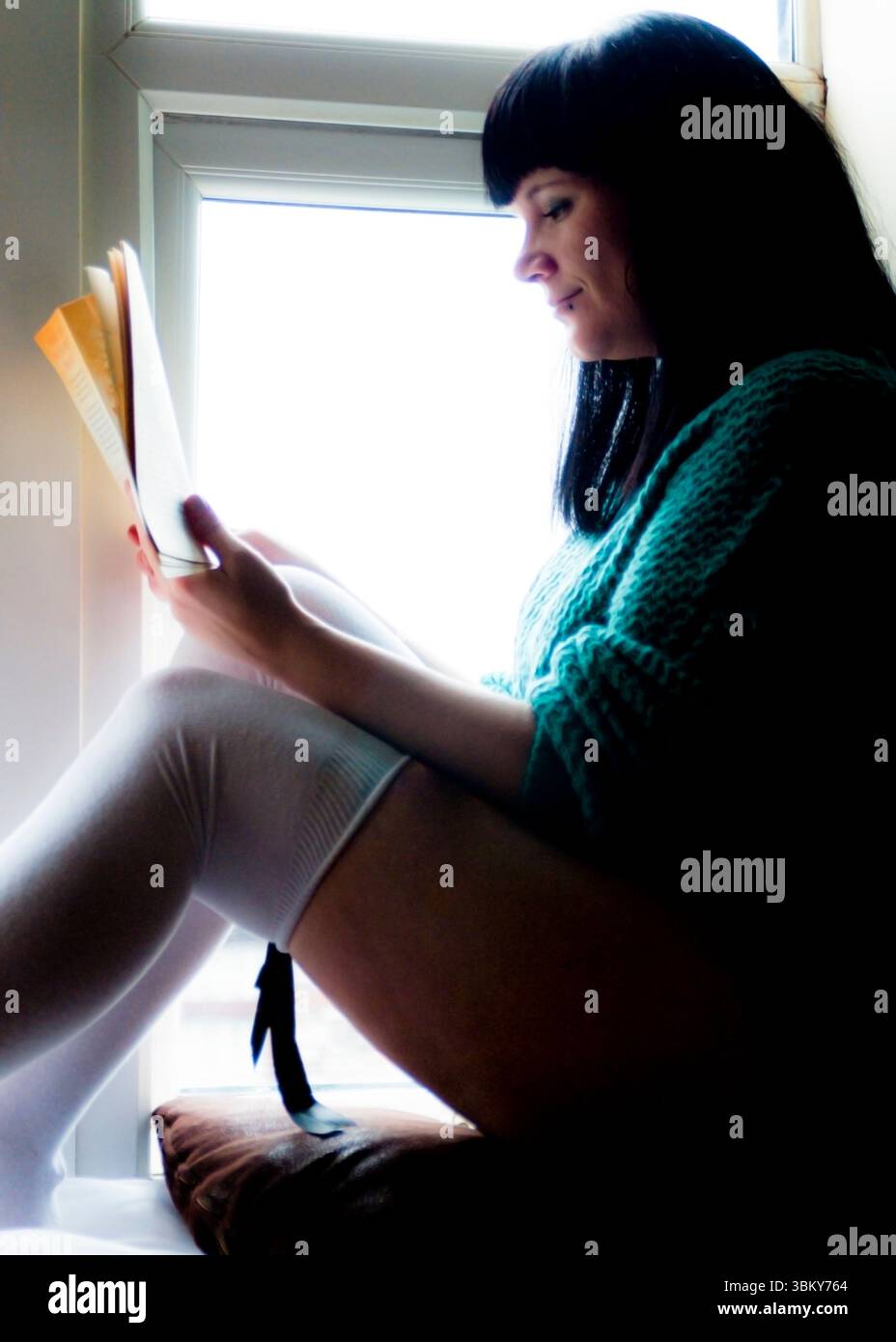 Une femme avec les cheveux noirs et un pull sarcelle est assise dans un siège de fenêtre, lisant un livre. Banque D'Images