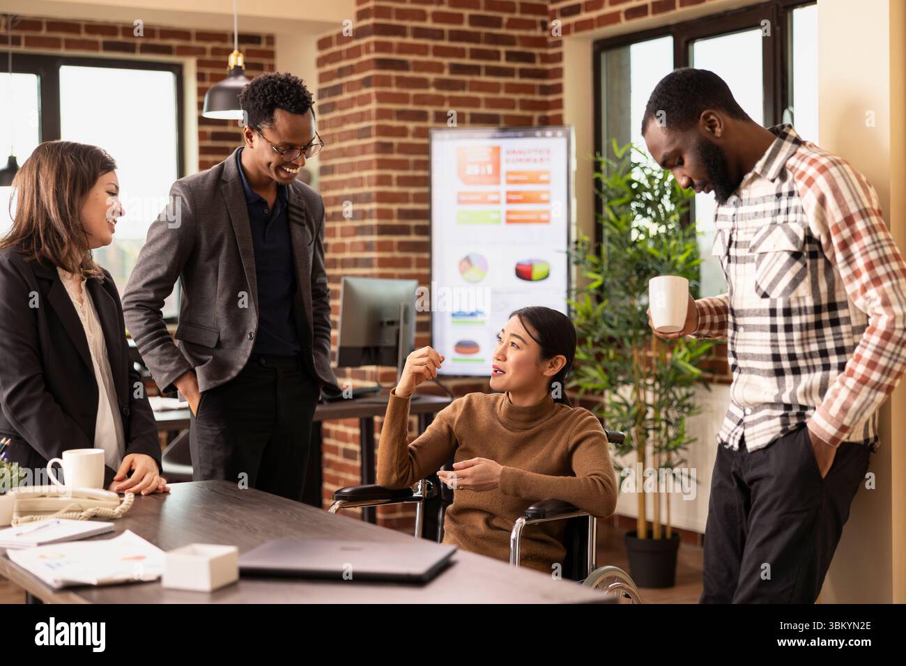 Équipe de start-up multiethnique debout et écoutant leur collègue en fauteuil roulant alors qu’elle partage une histoire hilarante. Groupe diversifié d'employés se liant pendant leur pause dans un espace de travail moderne. Banque D'Images