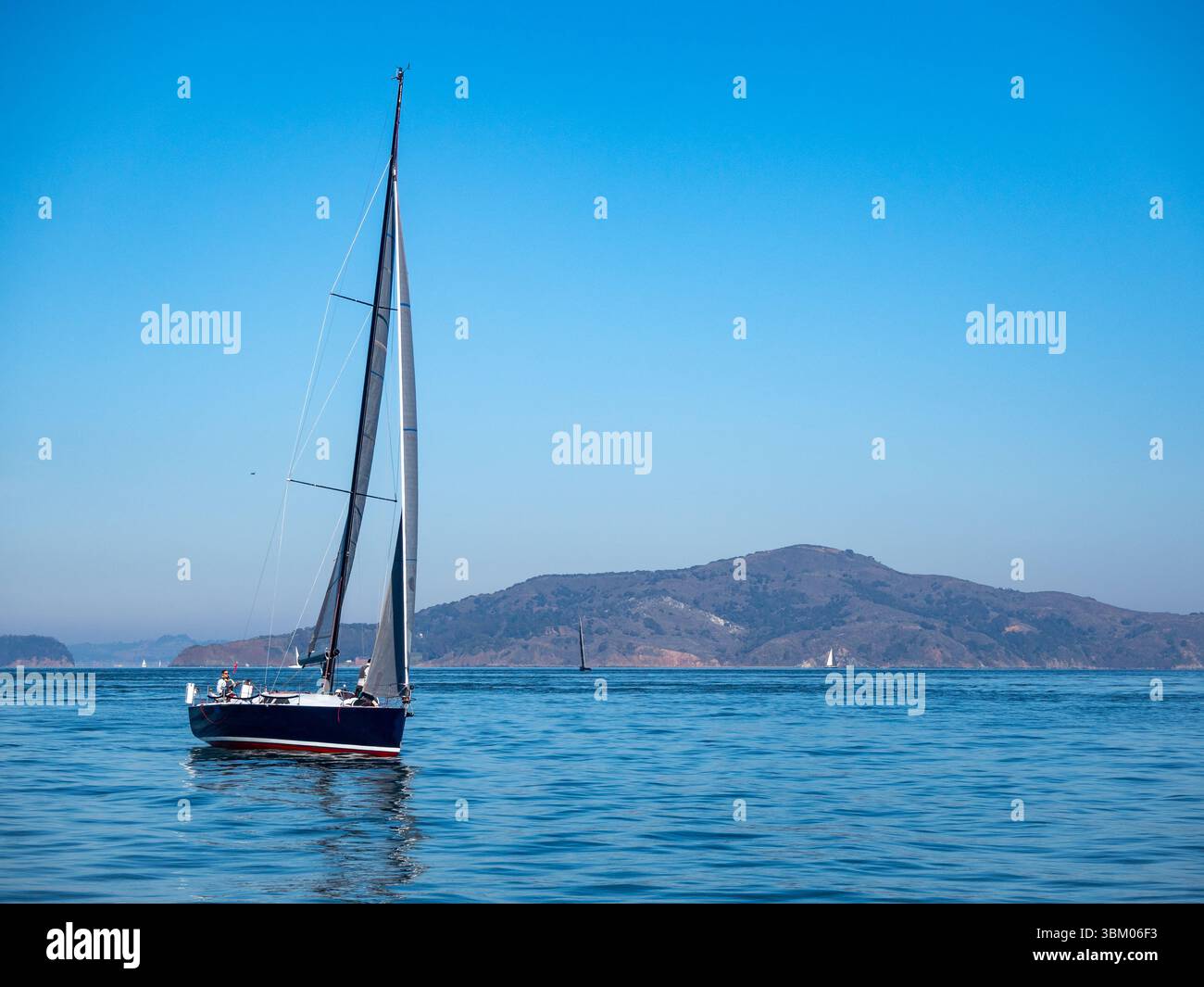 Un voilier navigue sur l'eau par temps clair. Le bateau est bleu foncé avec une bande rouge et un mât haut. Il y a d'autres voiliers au loin, ainsi qu'une chaîne de montagnes. Banque D'Images Un voilier navigue sur l'eau par temps clair. Le bateau est bleu foncé avec une bande rouge et un mât haut. Il y a d'autres voiliers au loin, ainsi qu'une chaîne de montagnes. Banque D'Images