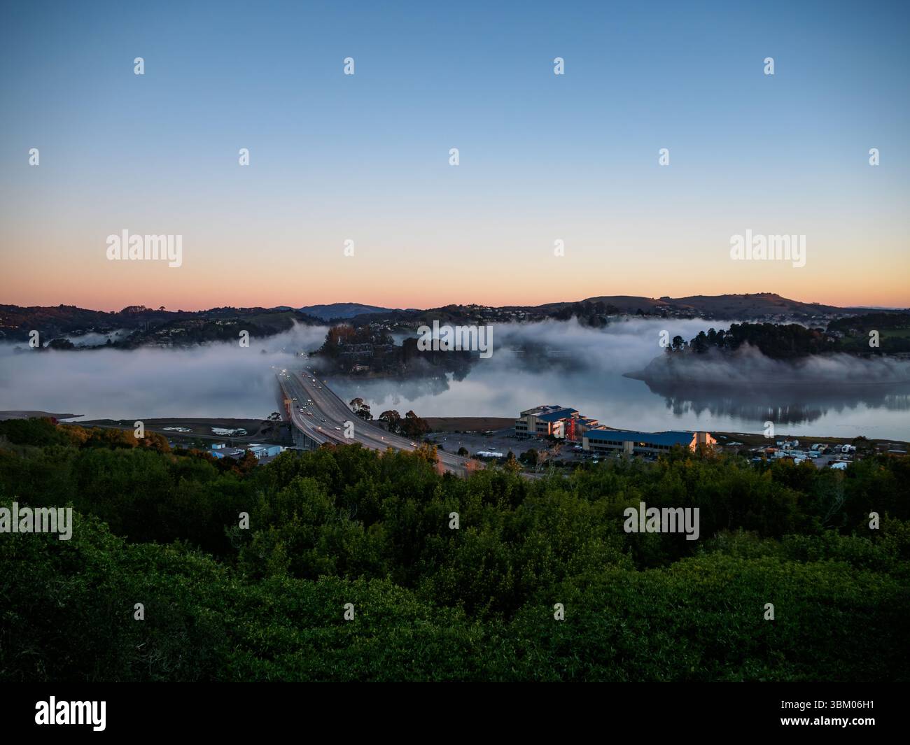Une vue aérienne d'un pont avec des voitures circulant dessus, entouré de brouillard et d'eau, avec des bâtiments et des arbres visibles en arrière-plan, pendant un lever ou coucher de soleil. Banque D'Images Une vue aérienne d'un pont avec des voitures circulant dessus, entouré de brouillard et d'eau, avec des bâtiments et des arbres visibles en arrière-plan, pendant un lever ou coucher de soleil. Banque D'Images
