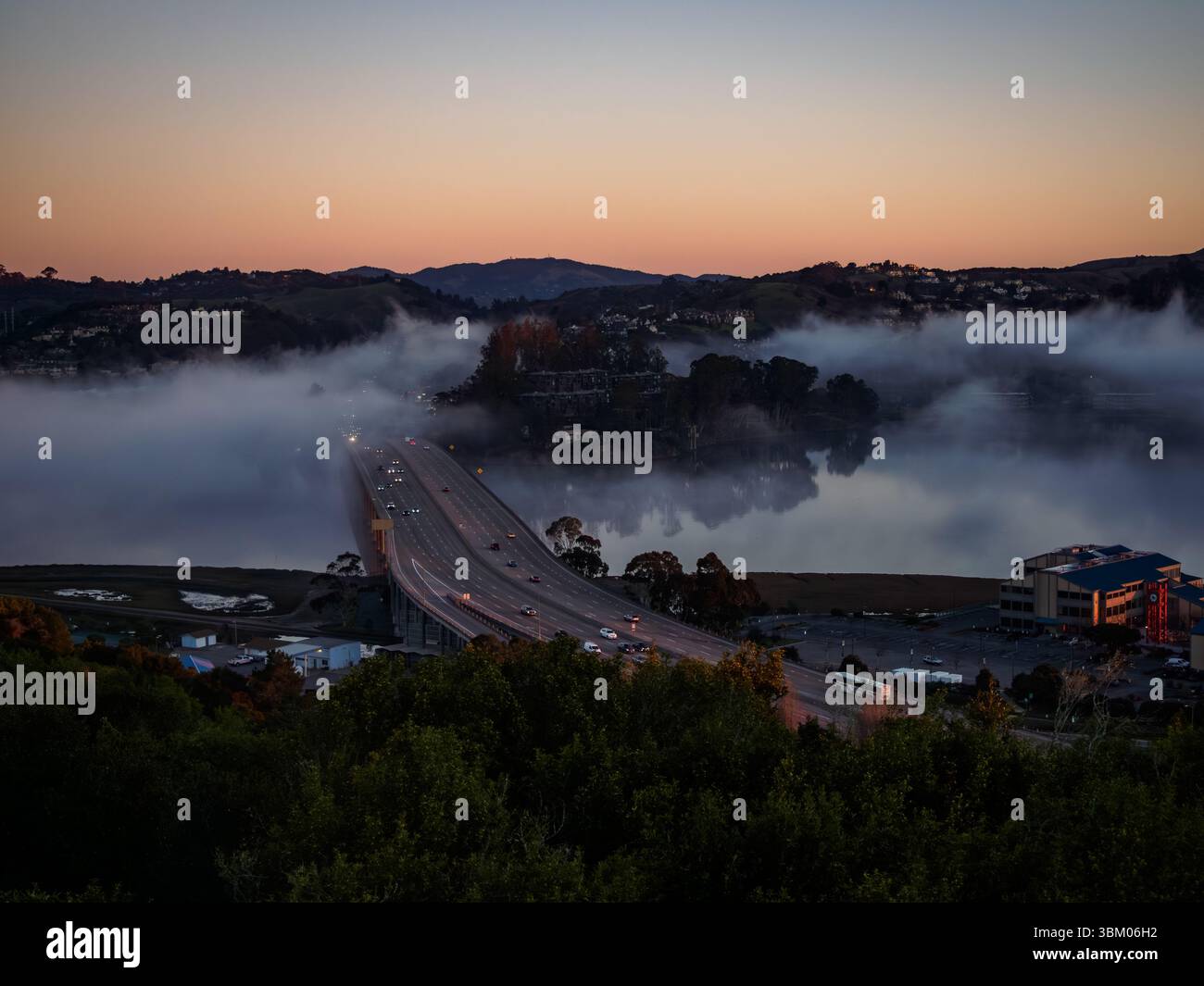 Les voitures traversent un pont entouré de brouillard au coucher du soleil, reliant les masses terrestres et facilitant le transport. Banque D'Images Les voitures traversent un pont entouré de brouillard au coucher du soleil, reliant les masses terrestres et facilitant le transport. Banque D'Images