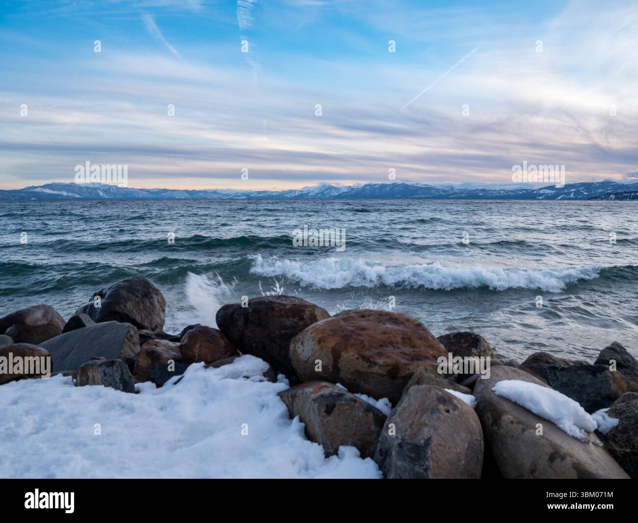 Les vagues s'écrasent contre des rochers enneigés sur la rive d'un lac, avec des montagnes visibles au loin sous un ciel nuageux. Banque D'Images Les vagues s'écrasent contre des rochers enneigés sur la rive d'un lac, avec des montagnes visibles au loin sous un ciel nuageux. Banque D'Images