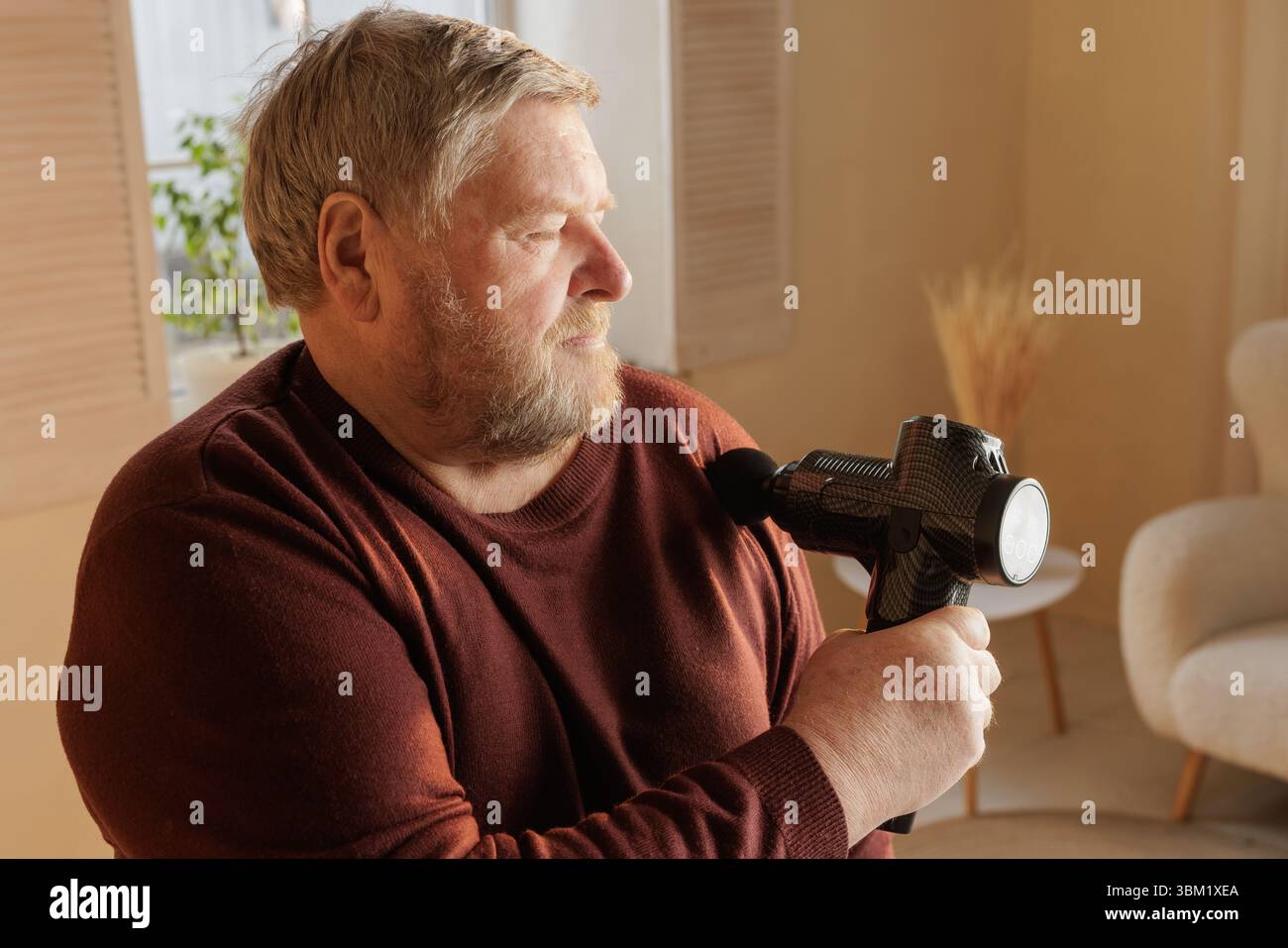 Homme âgé utilisant un pistolet de massage sur son épaule pour le soulagement de la douleur musculaire et l'auto-soin à la maison à la lumière naturelle. Banque D'Images