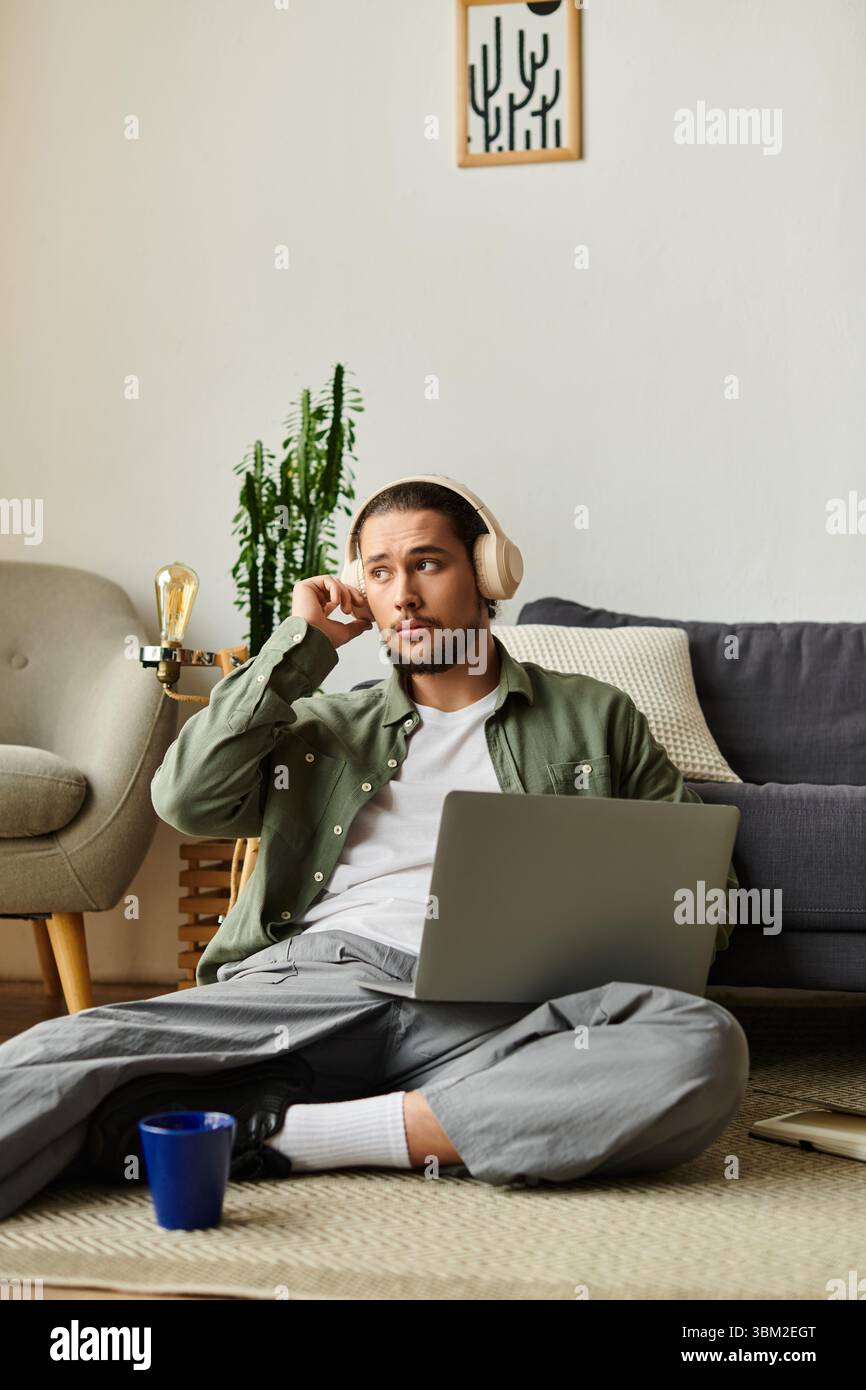 Un jeune homme est assis confortablement sur le sol à la maison, profondément dans la contemplation tout en utilisant son ordinateur portable. Banque D'Images
