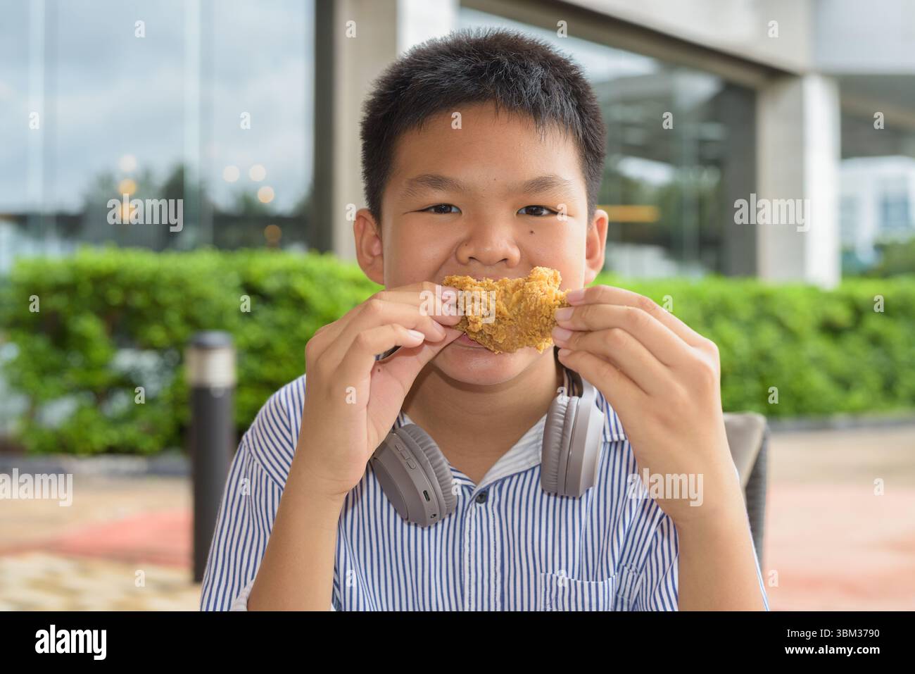 Garçon étudiant thaïlandais mangeant du poulet frit avec des écouteurs à la table extérieure Banque D'Images