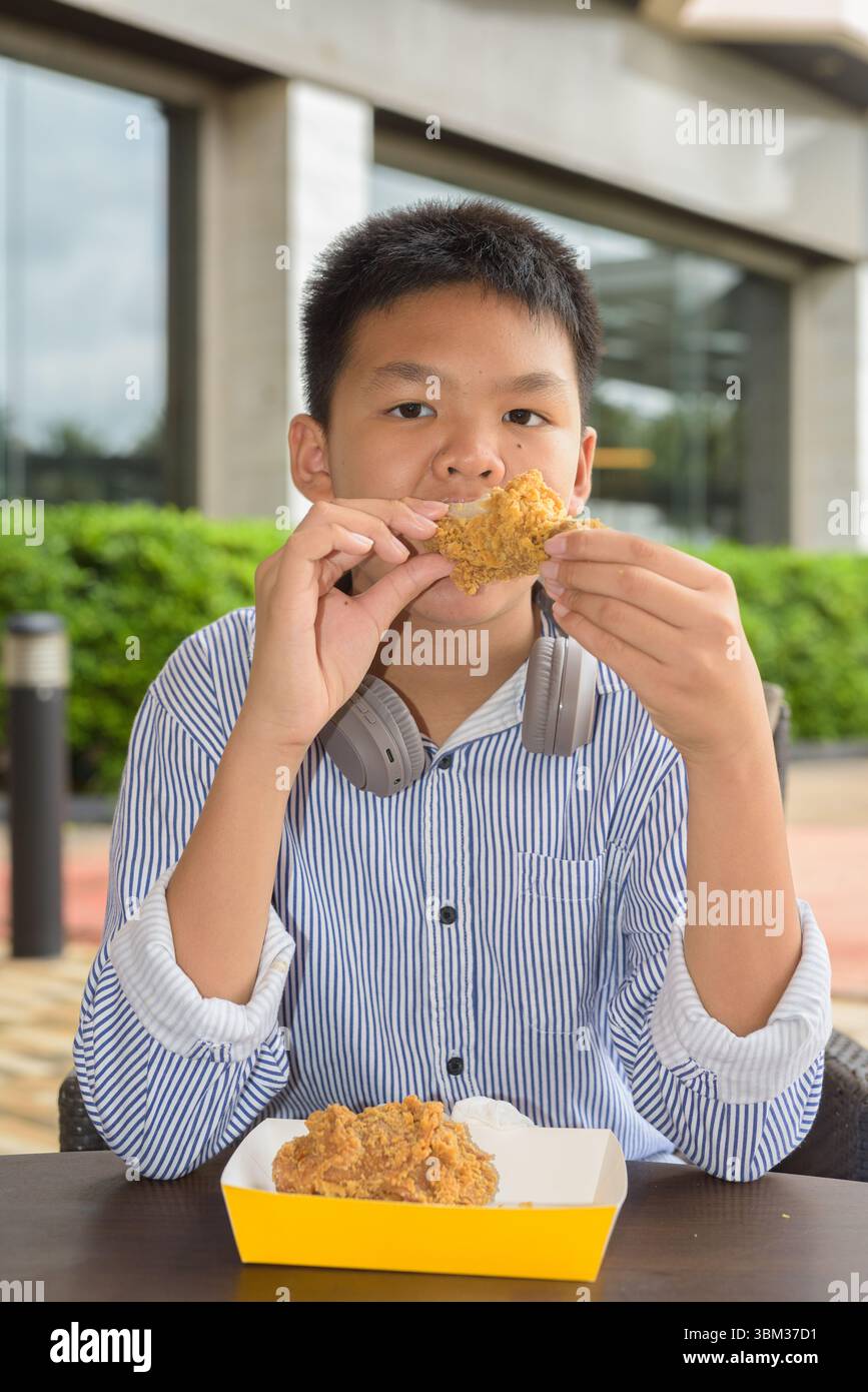 Garçon étudiant thaïlandais mangeant du poulet frit avec des écouteurs à la table extérieure Banque D'Images