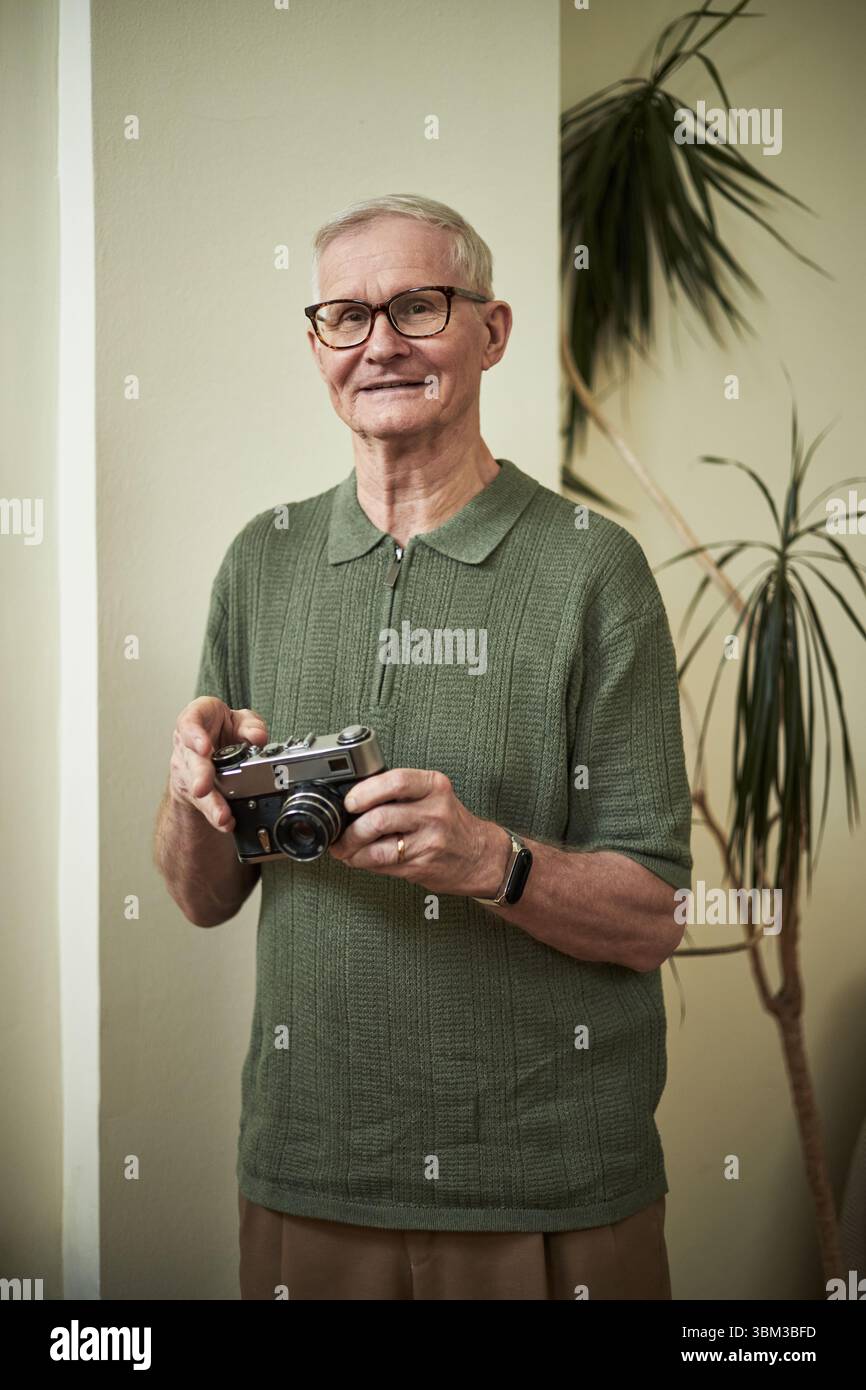 Homme âgé avec des lunettes souriant tout en tenant un appareil photo vintage, debout à l'intérieur devant une plante en pot, portant une chemise verte et regardant le contenu Banque D'Images