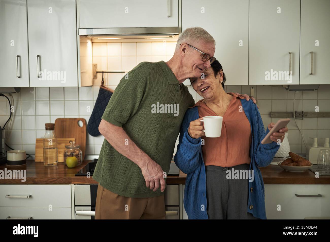 Couple âgé debout dans la cuisine, souriant tout en prenant le café du matin et en utilisant un smartphone. Dame senior tenant une tasse, homme senior embrassant affectueusement Banque D'Images