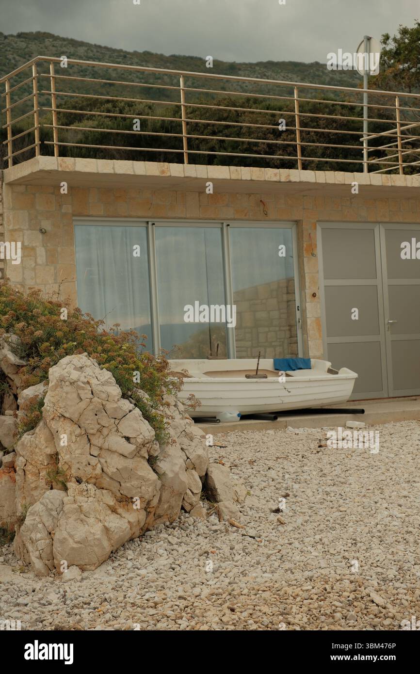 Un petit bâtiment en bord de mer avec des murs en pierre et des portes coulissantes en verre, une barque blanche reposant sur le sol en face. Banque D'Images