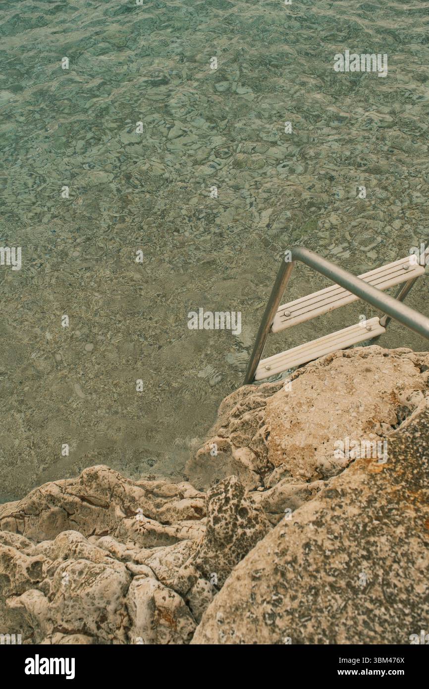 Gros plan sur un littoral rocheux avec du calcaire texturé et une échelle en acier inoxydable descendant dans une eau turquoise incroyablement claire. Banque D'Images