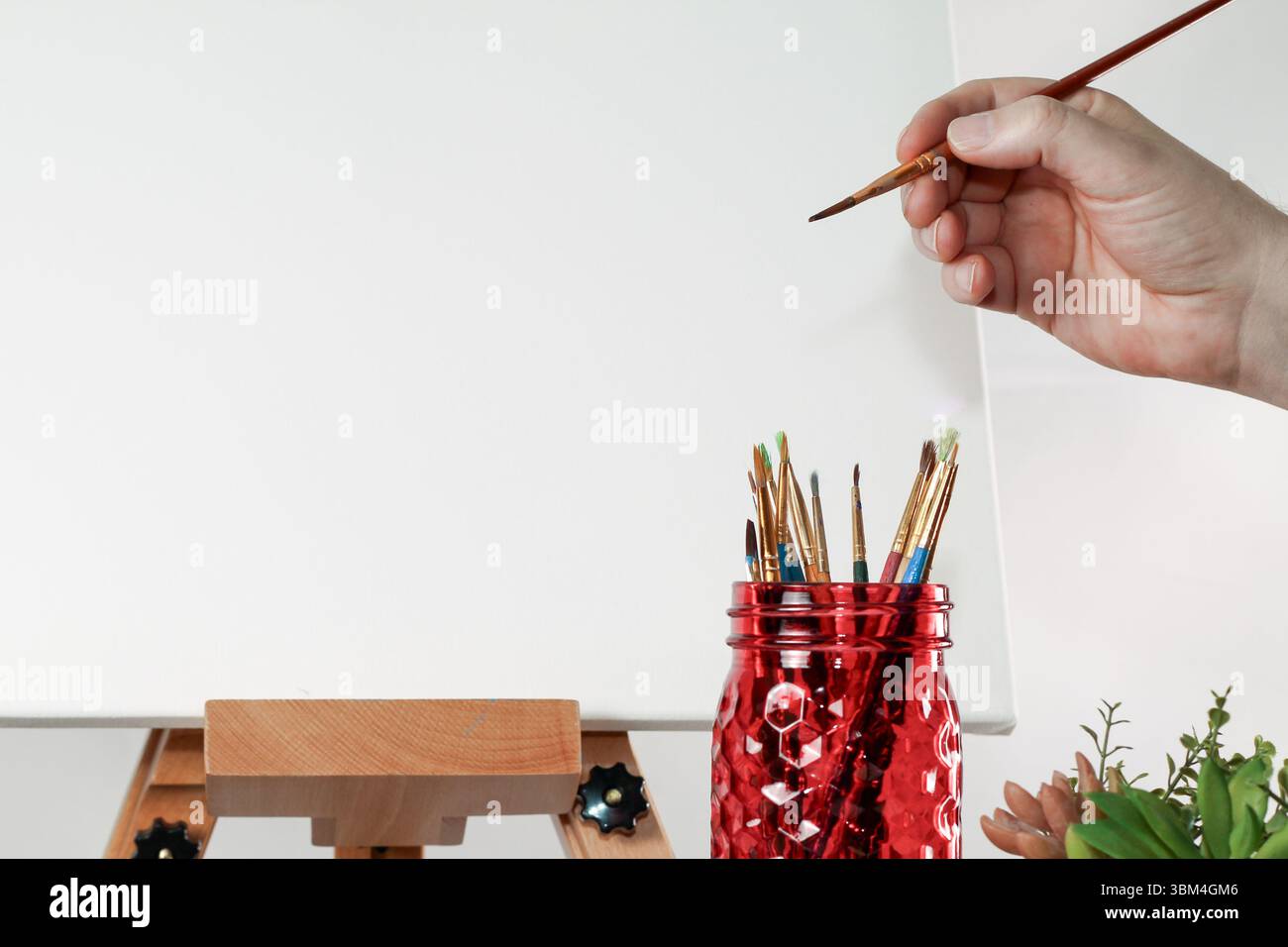 Main d’artiste tenant un pinceau à côté de Mason Jar et Canvas – scène Creative Workspace Banque D'Images Main d’artiste tenant un pinceau à côté de Mason Jar et Canvas – scène Creative Workspace Banque D'Images