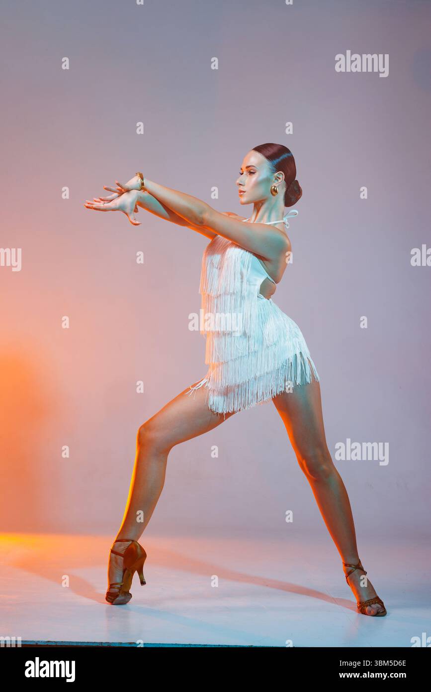 Danseuse latine dynamique en robe à franges blanches posant énergiquement en studio. Performance de danse latino-américaine moderne, élégante et élégante. Banque D'Images