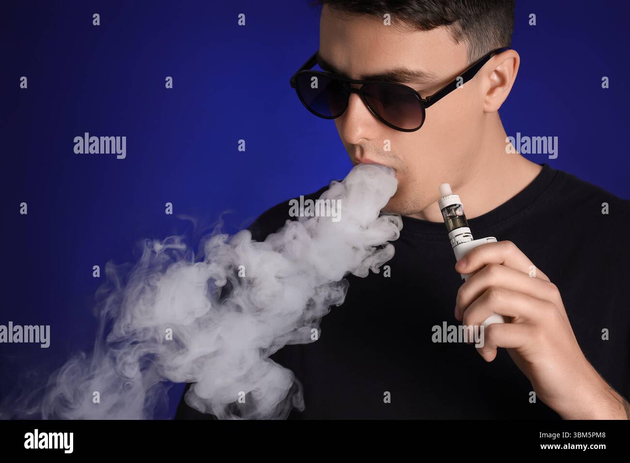 Jeune homme dans des lunettes de soleil fumant cigarette électronique sur fond bleu foncé, gros plan Banque D'Images