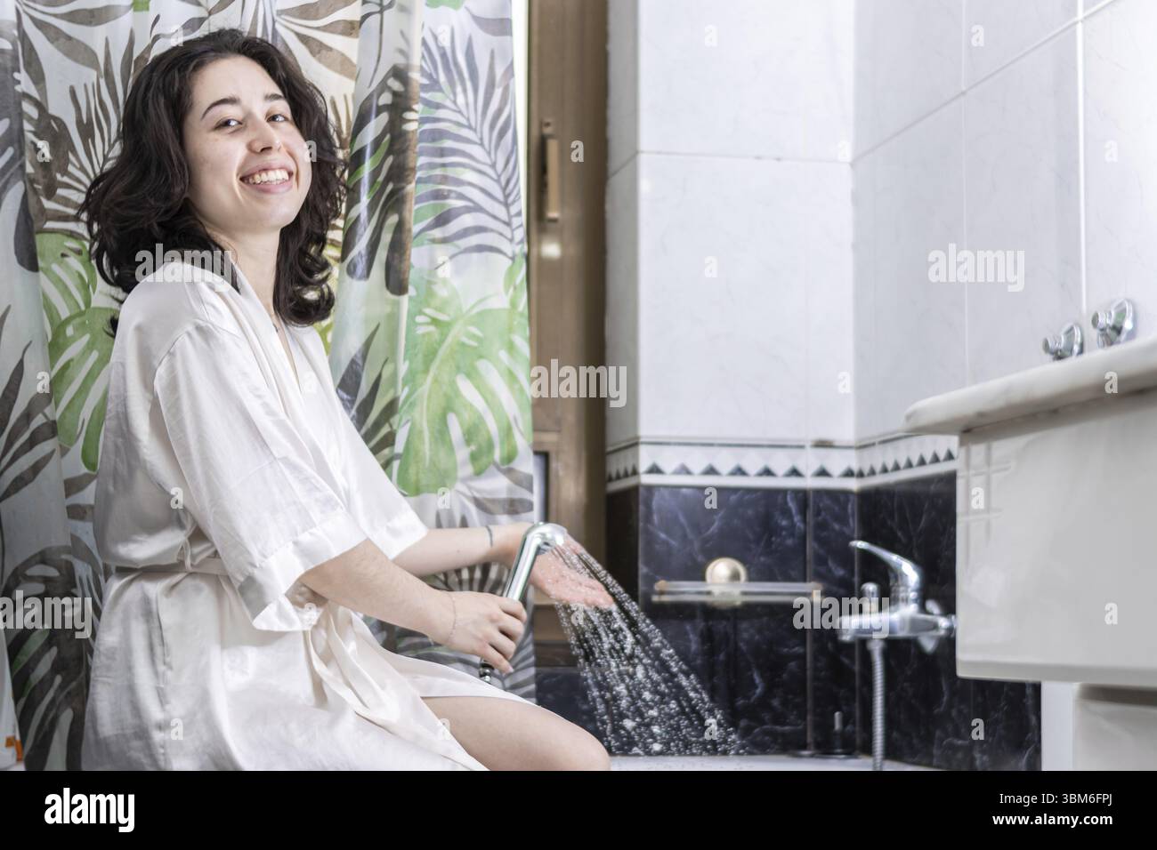 Jolie femme souriante portant un peignoir assis sur le bord de la baignoire remplissant d'eau Banque D'Images