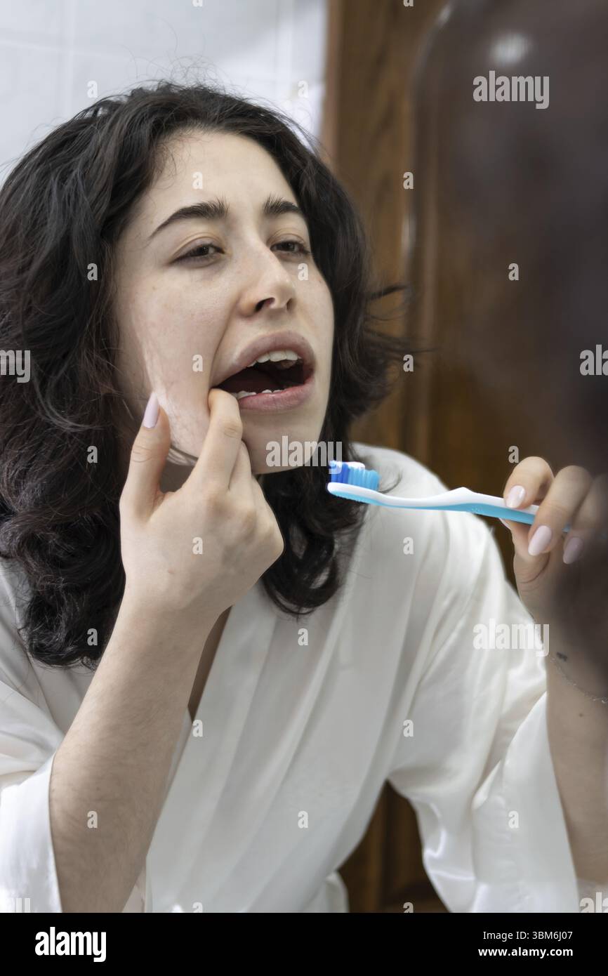 Portrait d'une femme latine tenant une brosse à dents regardant ses molaires et ses dents dans le miroir. les problèmes d'hygiène buccale chez les jeunes sont très fréquents Banque D'Images