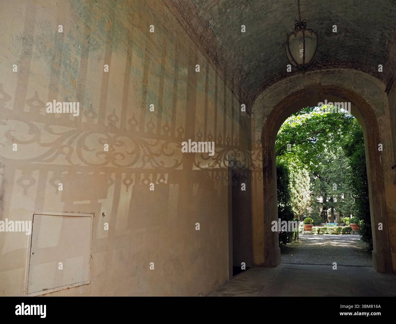 Des ombres de portes en ferronnerie ornées projetées par la lumière du soleil sur le mur latéral de l'arche plâtrée de l'entrée d'une cour verdoyante à Florence, en Italie Banque D'Images