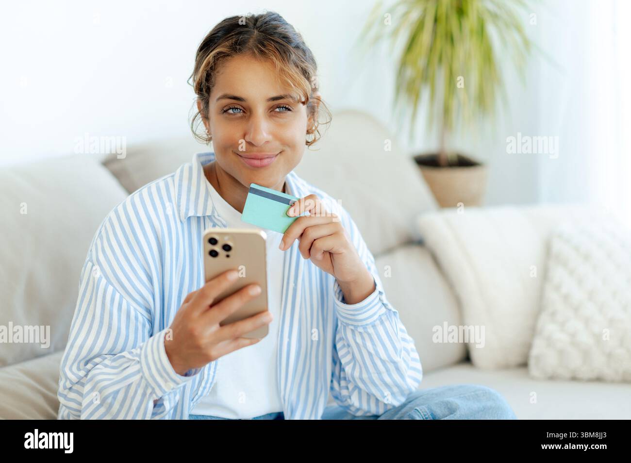 Femme brésilienne positive tenant une carte de crédit et un smartphone, faisant l'achat en ligne ou la banque à la maison assis au canapé. Elle a l'air heureuse et réfléchie tout en magasinant ou en gérant les finances sur mobile Banque D'Images