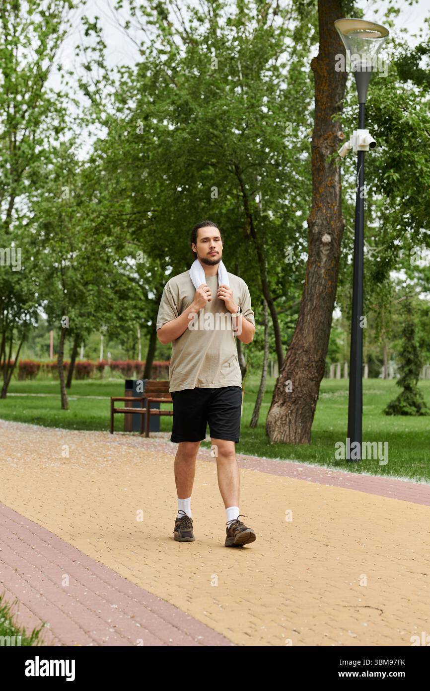 Un jeune homme en forme avec une barbe marche en toute confiance à travers un chemin de parc paisible, faisant de l'exercice en plein air. Banque D'Images