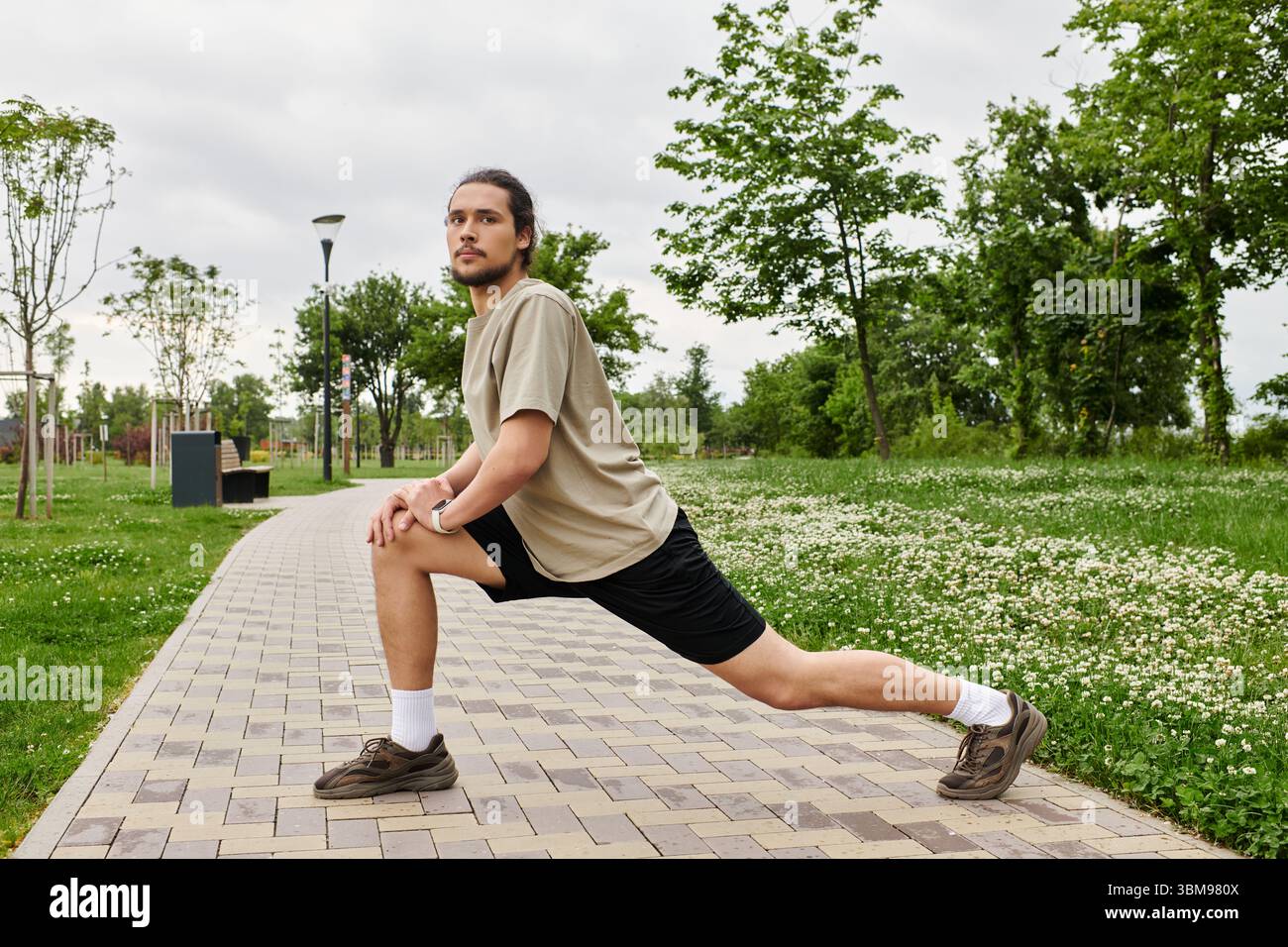 Homme sportif avec la barbe s'étire par le chemin dans un parc verdoyant pour booster ses performances athlétiques. Banque D'Images