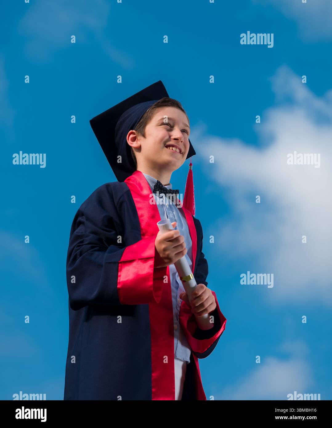 Beau garçon avec tenue de graduation d'école primaire et diplôme. Garçon dans des robes de graduation regardant haut avec fond de ciel Banque D'Images