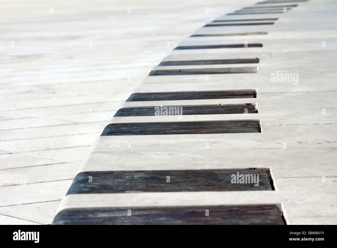 Banc en béton incurvé avec des lattes en bois sombre espacées uniformément placées dans un trottoir blanc, formant un motif géométrique répétitif dans une place urbaine ensoleillée Banque D'Images
