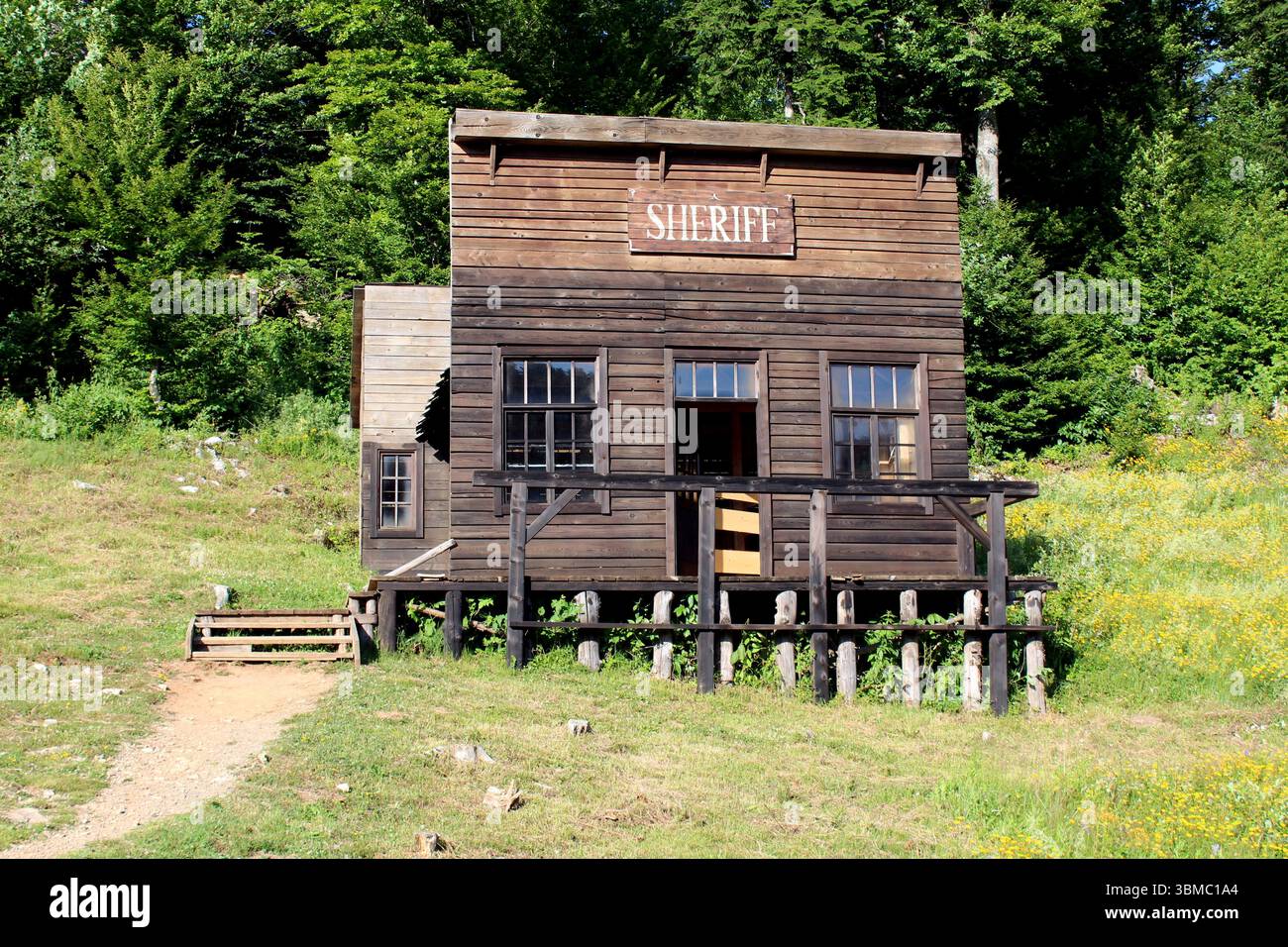 Vieux bureau de shérifs en bois avec porche et panneaux se dresse sur des poteaux dans une clairière herbeuse, entouré de fleurs sauvages et d'une forêt verte dense sous-jacente Banque D'Images