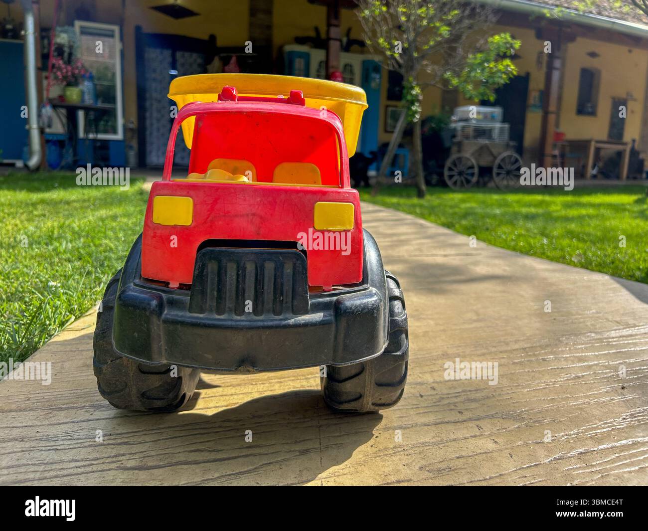 Camion à benne en plastique rouge et jaune placé sur un chemin en béton dans une cour ensoleillée avec de l'herbe et une maison rustique en arrière-plan. - Image de stock capturée avec un smartphone