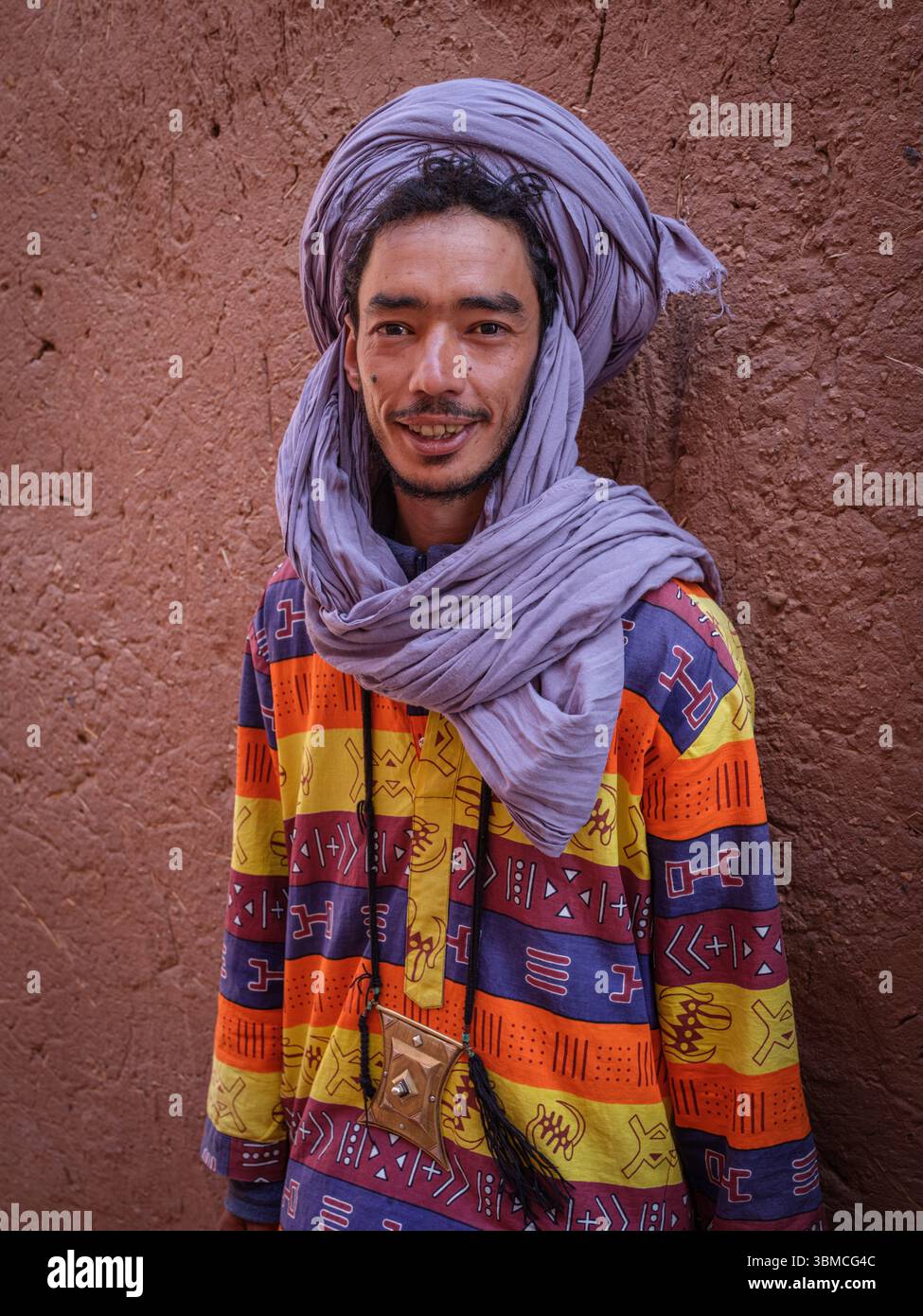 Maroc, 25 mai 2025. Portrait d'un jeune homme amazigh (berbère) souriant portant une tenue traditionnelle et un turban, debout contre un mur de terre dans un Banque D'Images