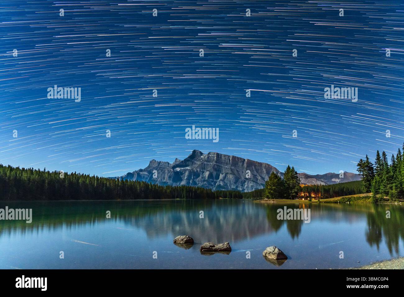 Les sentiers étoilés au-dessus du mont Rundle depuis Two Jack Lake la nuit, avec le ciel étoilé et la montagne reflétés dans l'eau. Parc national Banff, Alberta, Canada. Banque D'Images