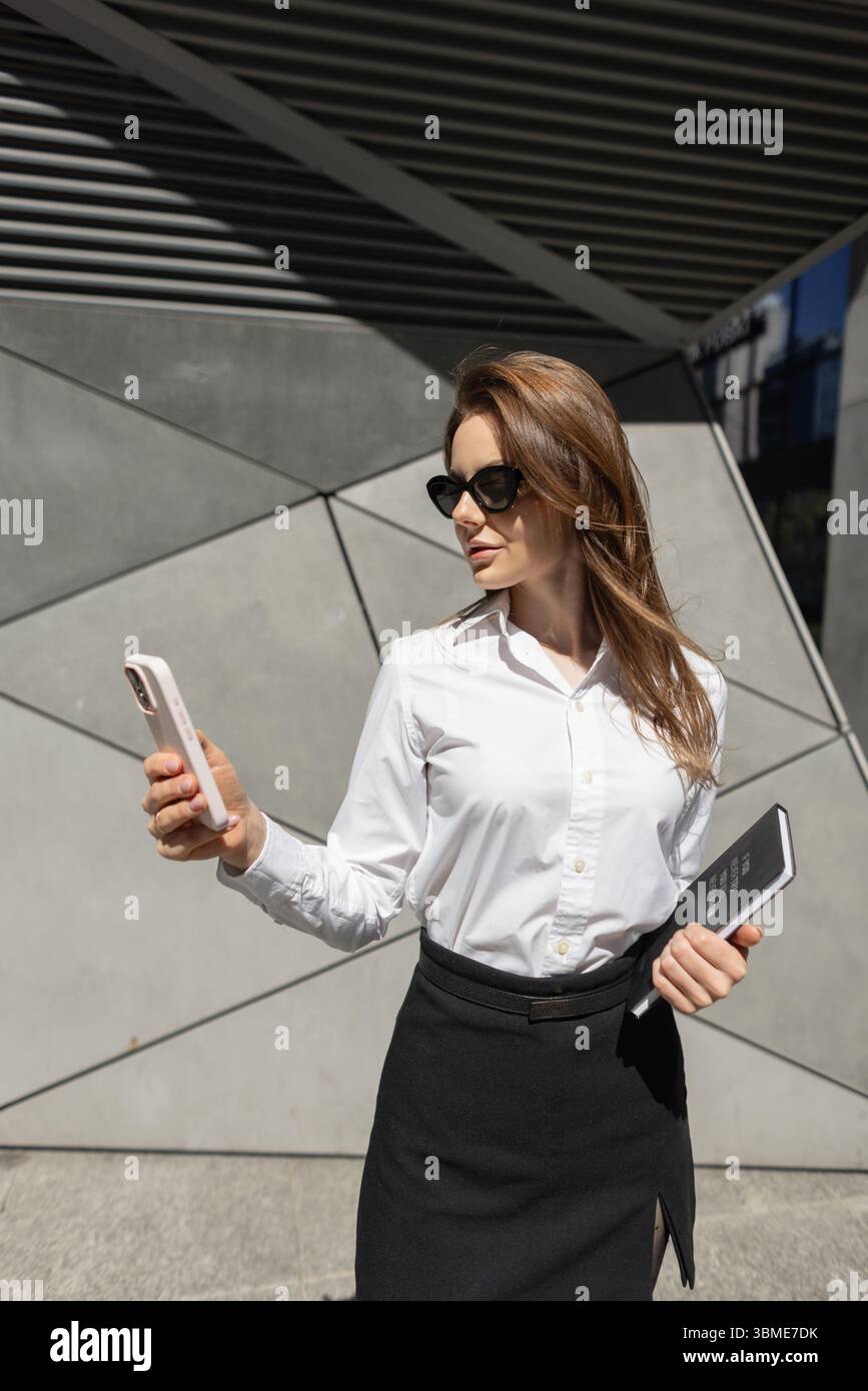 Jeune femme d'affaires dans des lunettes de soleil vérifiant son téléphone tout en se tenant debout à l'extérieur D'Un immeuble de bureaux moderne. Confiant, élégant et connecté dans la ville. Banque D'Images