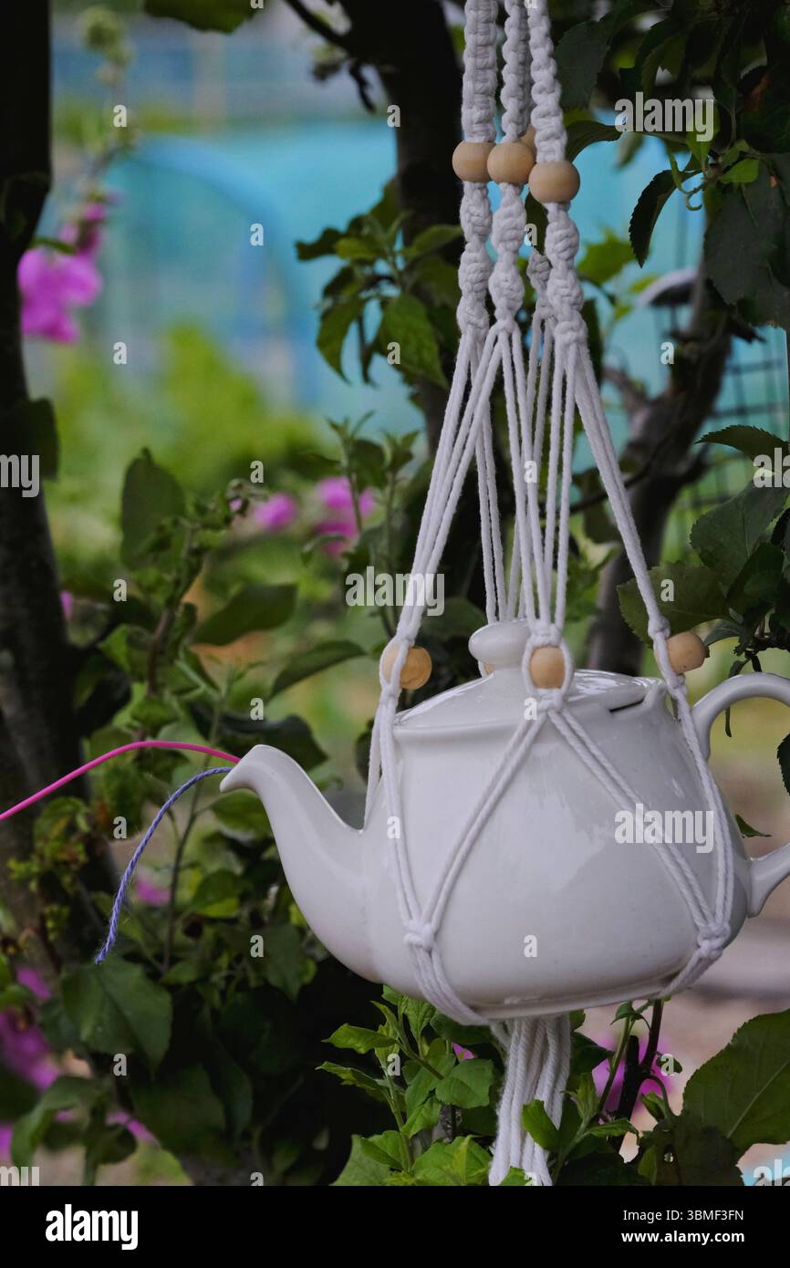 Creative allotissement jardinage hack — une théière en céramique blanche réutilisée comme distributeur de ficelle, suspendue à un arbre dans un porte-macramé fait main Banque D'Images