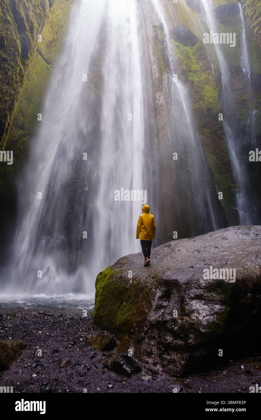 Une personne vêtue d'une veste de pluie jaune vif se tient sur un rocher, contemplant avec admiration la puissante cascade de Gljufrabui en Islande Banque D'Images
