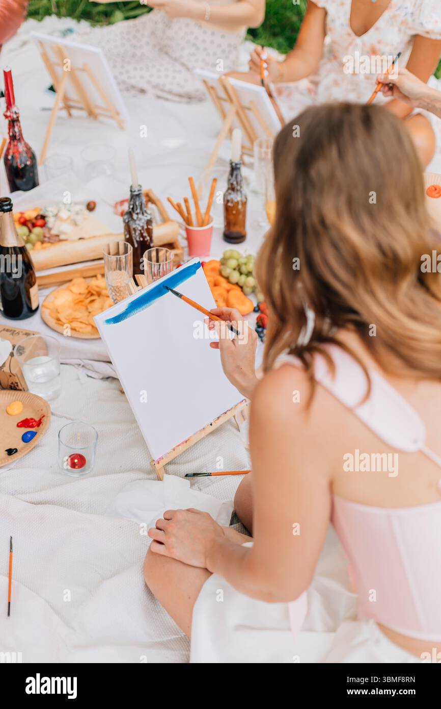 Femme en haut rose clair peignant ciel bleu sur toile blanche lors d'une séance de pique-nique artistique en plein air détendue. Banque D'Images