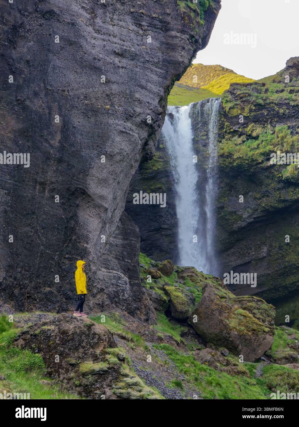 Une femme dans une veste de pluie jaune éclatante contraste magnifiquement avec les falaises rocheuses et la cascade de Kvernufoss. Femme en veste de pluie en Islande Banque D'Images