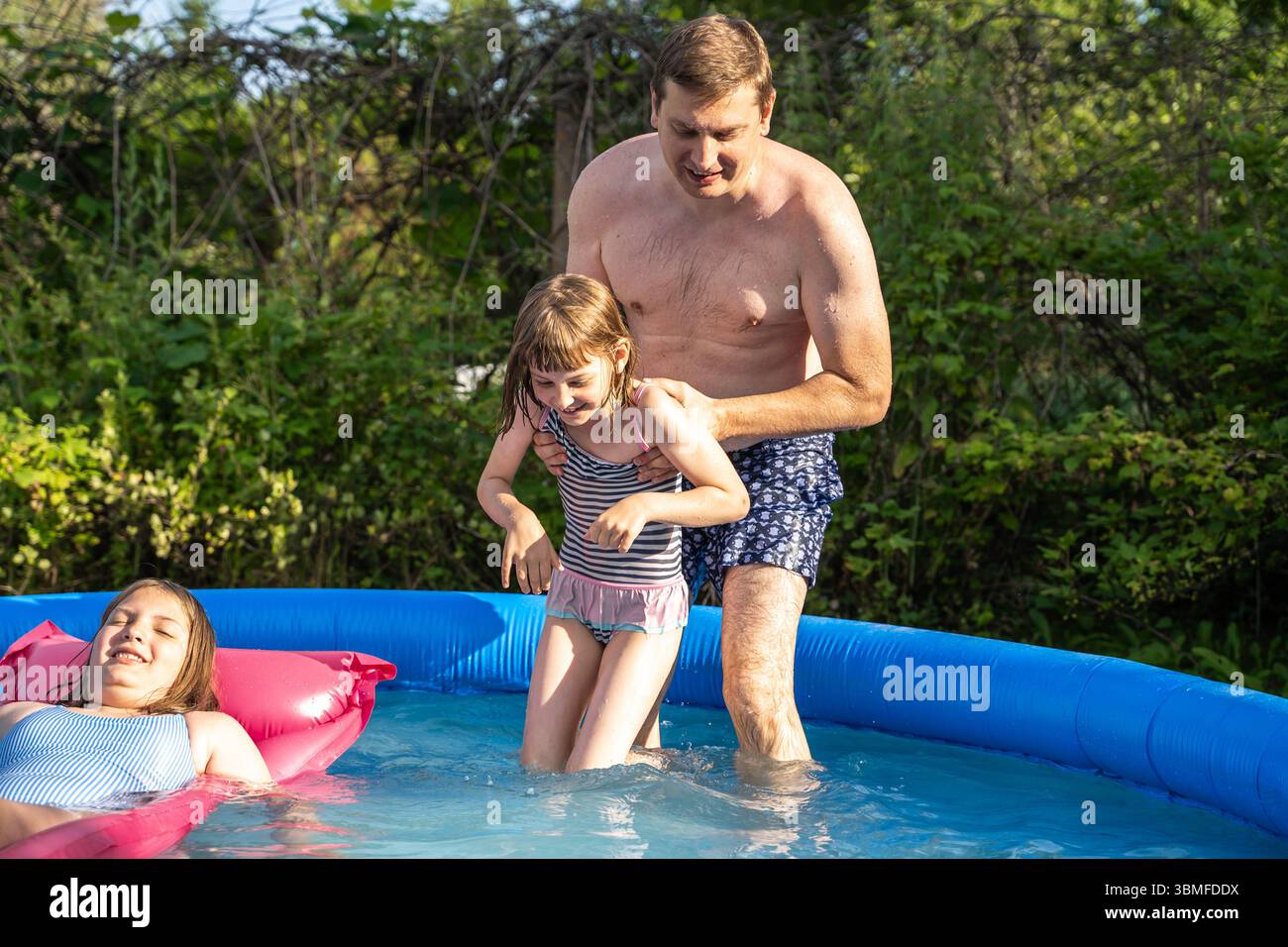Enfants jouant dans la piscine gonflable le jour d'été. Deux filles riant, éclaboussant de l'eau avec père. Amusement en famille, natation dans la cour, activité de plein air, b Banque D'Images