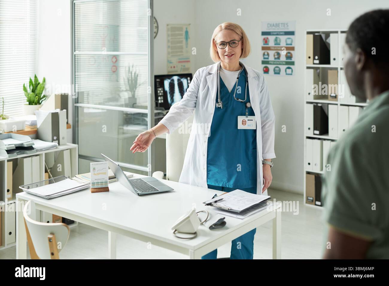 Médecin caucasien d'âge moyen accueillant un patient homme noir d'âge moyen dans le cabinet médical, faisant un geste vers le bureau avec des documents médicaux et digi Banque D'Images