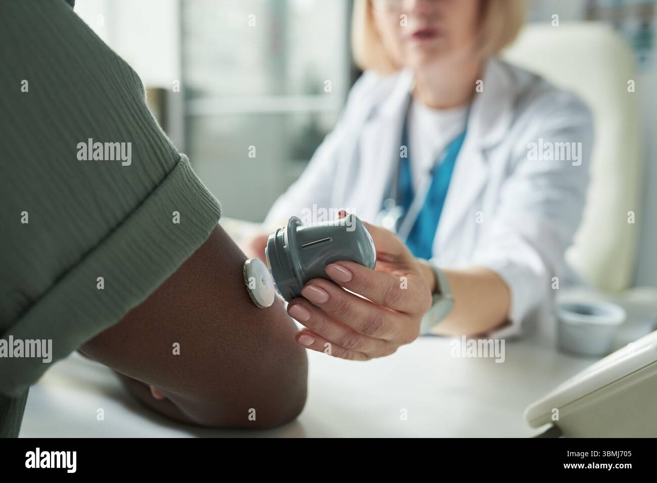 Médecin féminin caucasien d'âge moyen examinant le bras de l'homme noir à l'aide d'un dispositif de surveillance du glucose lors d'une consultation de diabète au cabinet médical, focalisation Banque D'Images