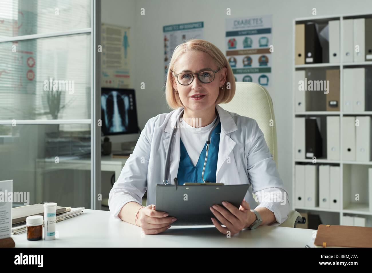 Portrait de femme caucasienne d'âge moyen médecin assis au bureau tenant une presse-papiers regardant dans la caméra dans le bureau médical avec des affiches de sensibilisation au diabète Banque D'Images