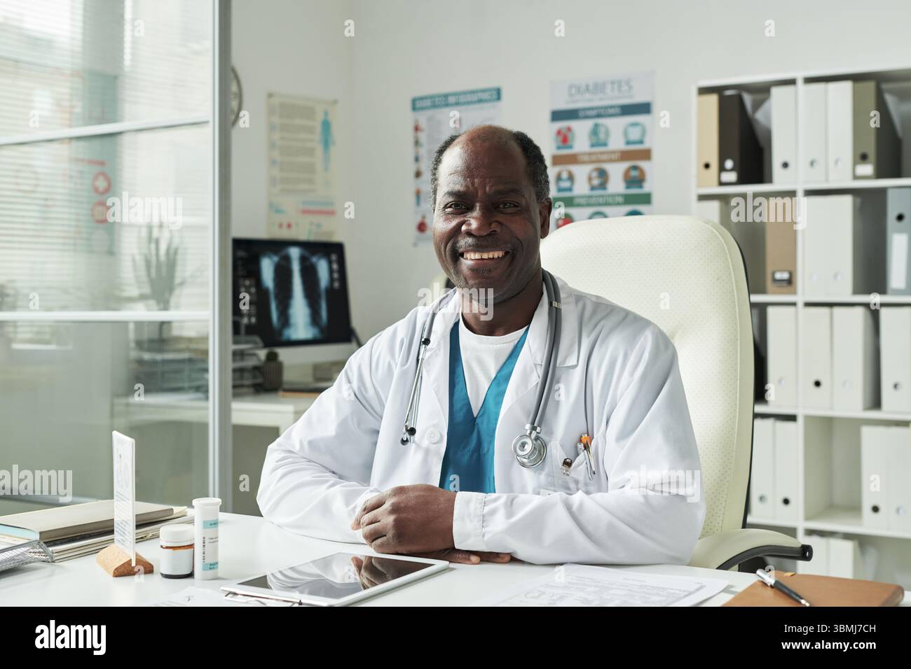 Portrait d'un médecin homme noir d'âge moyen assis au bureau dans le cabinet médical souriant à la caméra avec des affiches informatives sur le diabète en arrière-plan Banque D'Images
