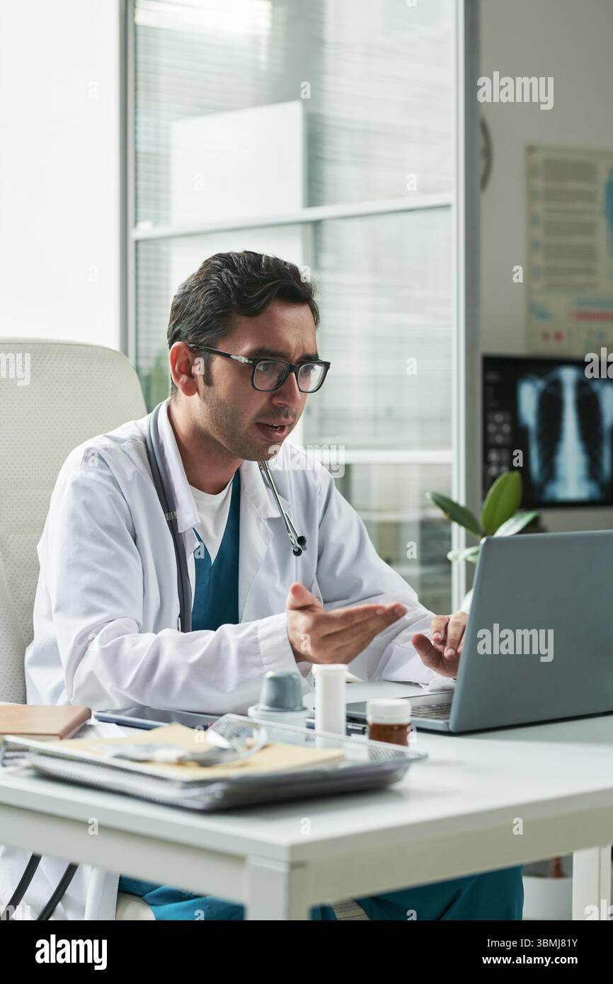 Médecin masculin caucasien d'âge moyen consultant le patient atteint de diabète par appel vidéo, faisant des gestes tout en étant assis au bureau avec ordinateur portable dans le cabinet médical, disque Banque D'Images