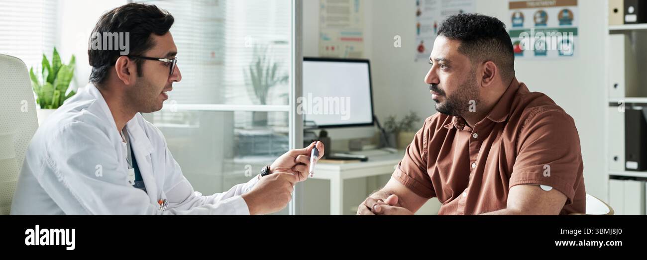 Homme d'Asie du Sud d'âge moyen assis dans le cabinet médical consultant avec un médecin masculin, discutant de la gestion du diabète tout en tenant le dispositif médical Banque D'Images