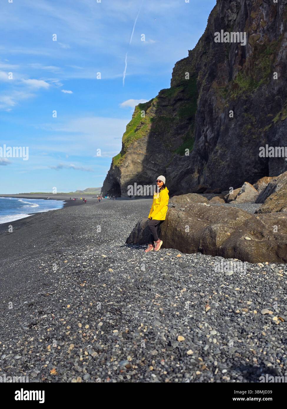 À Reynisfjara Beach, un voyageur se tient debout dans une veste de pluie jaune vif, entouré de falaises majestueuses et de sables noirs lisses. Les vagues calmes tapissent doucement sur le rivage sous un ciel bleu clair. Banque D'Images