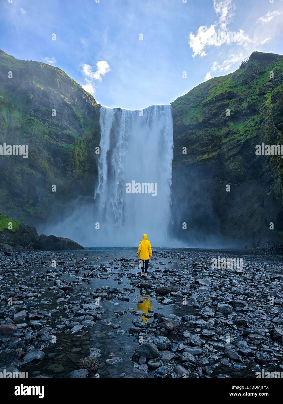 La cascade de Skogafoss descend en cascade les falaises majestueuses d'Islande, créant une atmosphère mystique. Femme en veste de pluie jaune debout près d'une cascade Banque D'Images