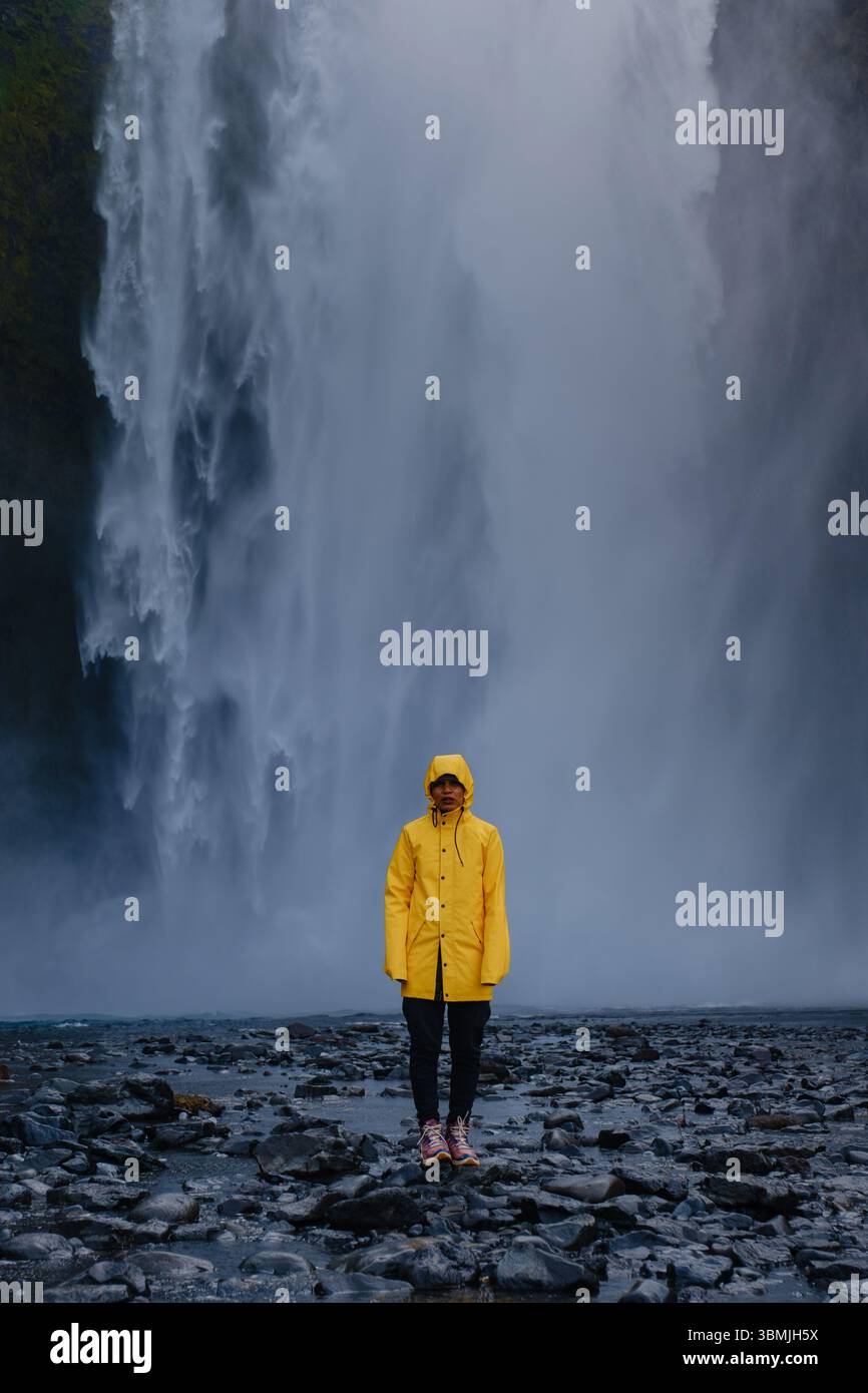 Une femme debout devant la majestueuse cascade de Skogafoss, un voyageur dans une veste de pluie jaune vif est entouré de brume et d'une beauté naturelle étonnante. Banque D'Images