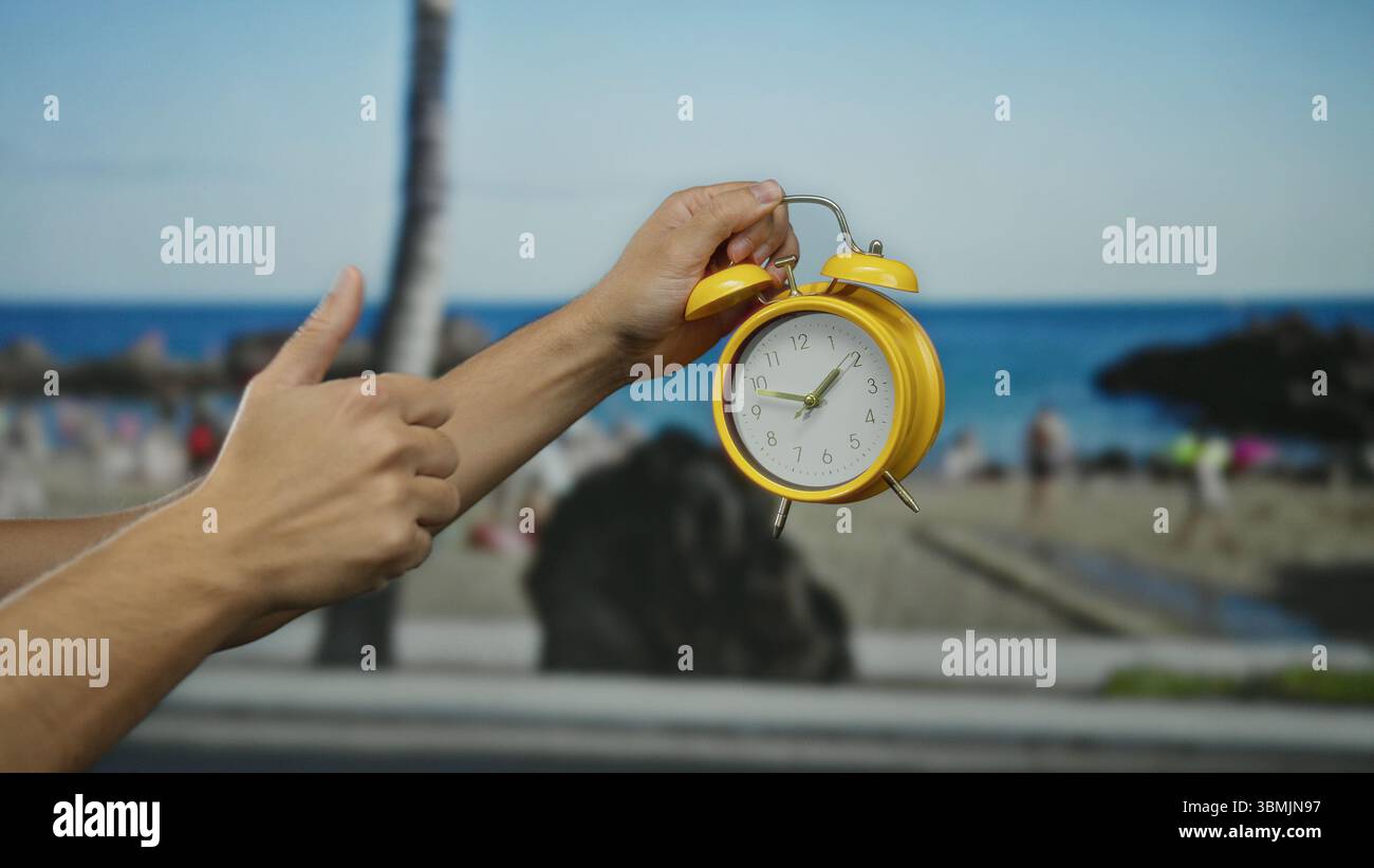 Main masculine tenant un réveil jaune à la plage avec fond flou de bord de mer sous le ciel bleu indiquant le concept de temps dans une atmosphère côtière détendue. Banque D'Images