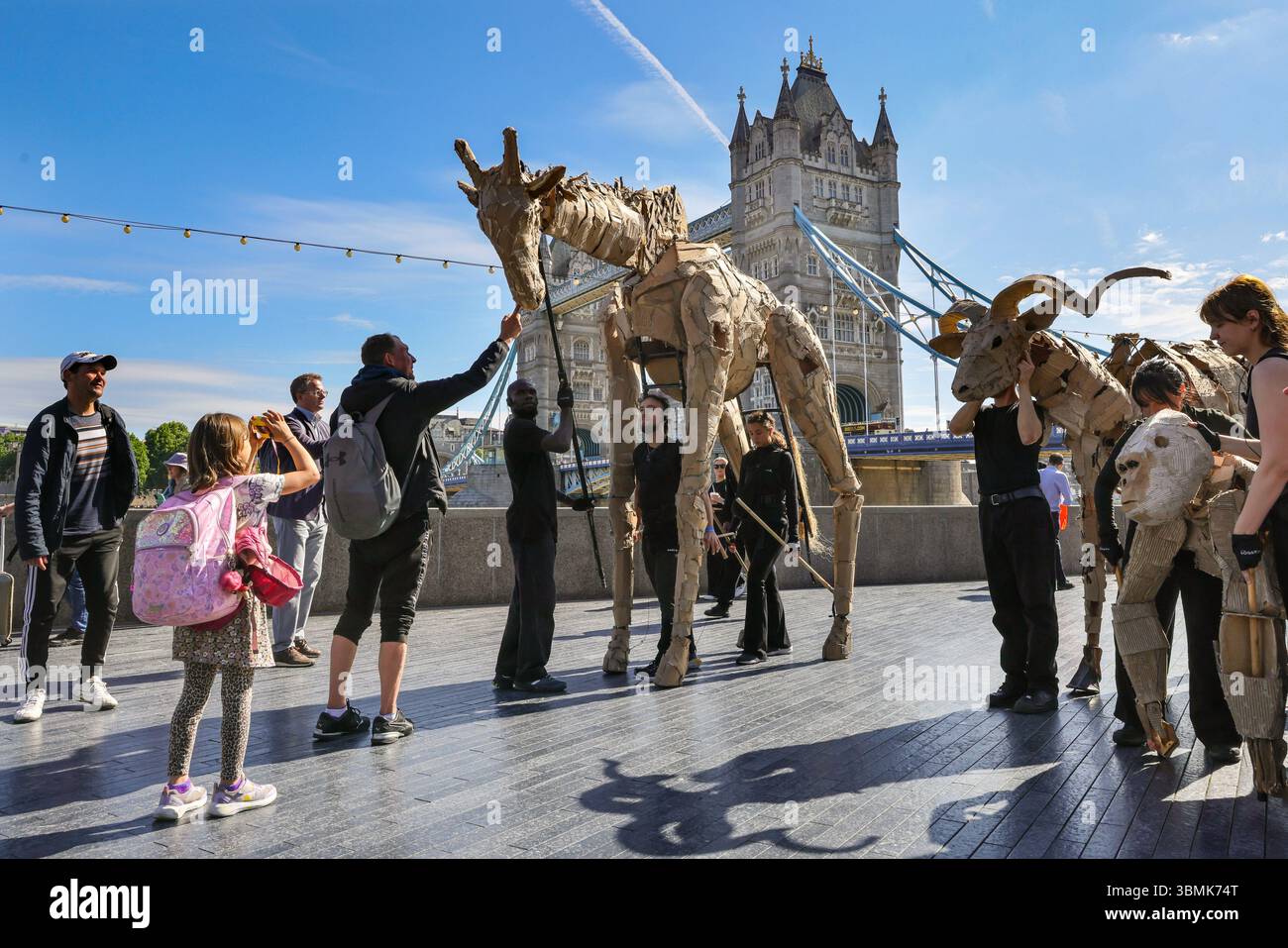 Londres, Royaume-Uni. 27 juin 2025. Les artistes marchent du Bridge Theatre à la Tamise avec Tower Bridge et la City de Londres derrière. LES TROUPEAUX sont l'art public et l'action climatique. D’avril à août 2025, les animaux marionnettes grandeur nature parcourent un itinéraire de 20 000 km entre le bassin du Congo et le cercle arctique, symbolisant leur fuite de la catastrophe climatique avec des arrêts dans de nombreuses villes et pays, à Londres du 27 au 29 juillet. Crédit : Imageplotter/Alamy Live News Banque D'Images