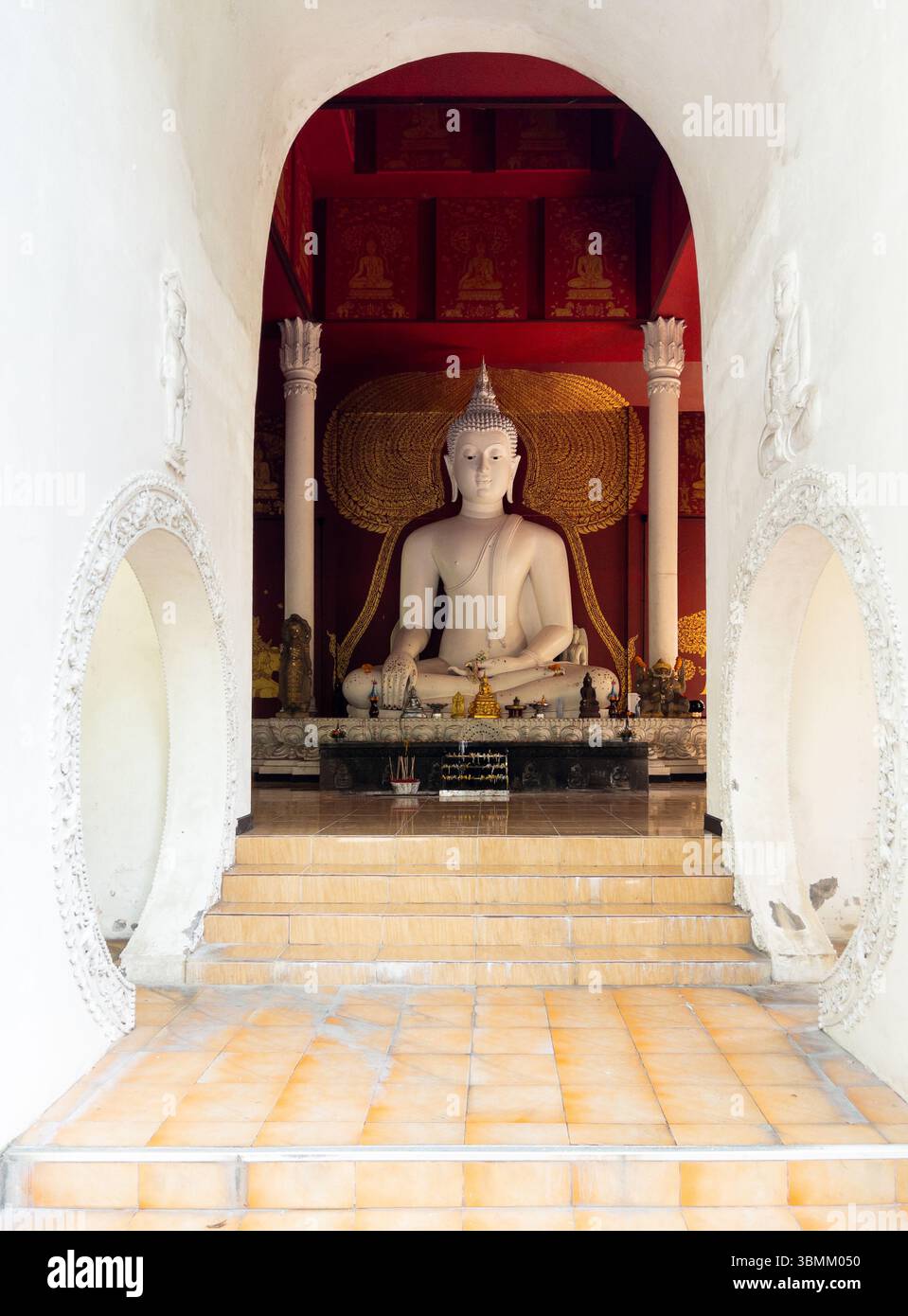 Une majestueuse statue blanche de Bouddha rayonne de tranquillité dans un temple sacré, encadré par des arches ornées et des accents dorés. L'intérieur serein invite l'esprit Banque D'Images