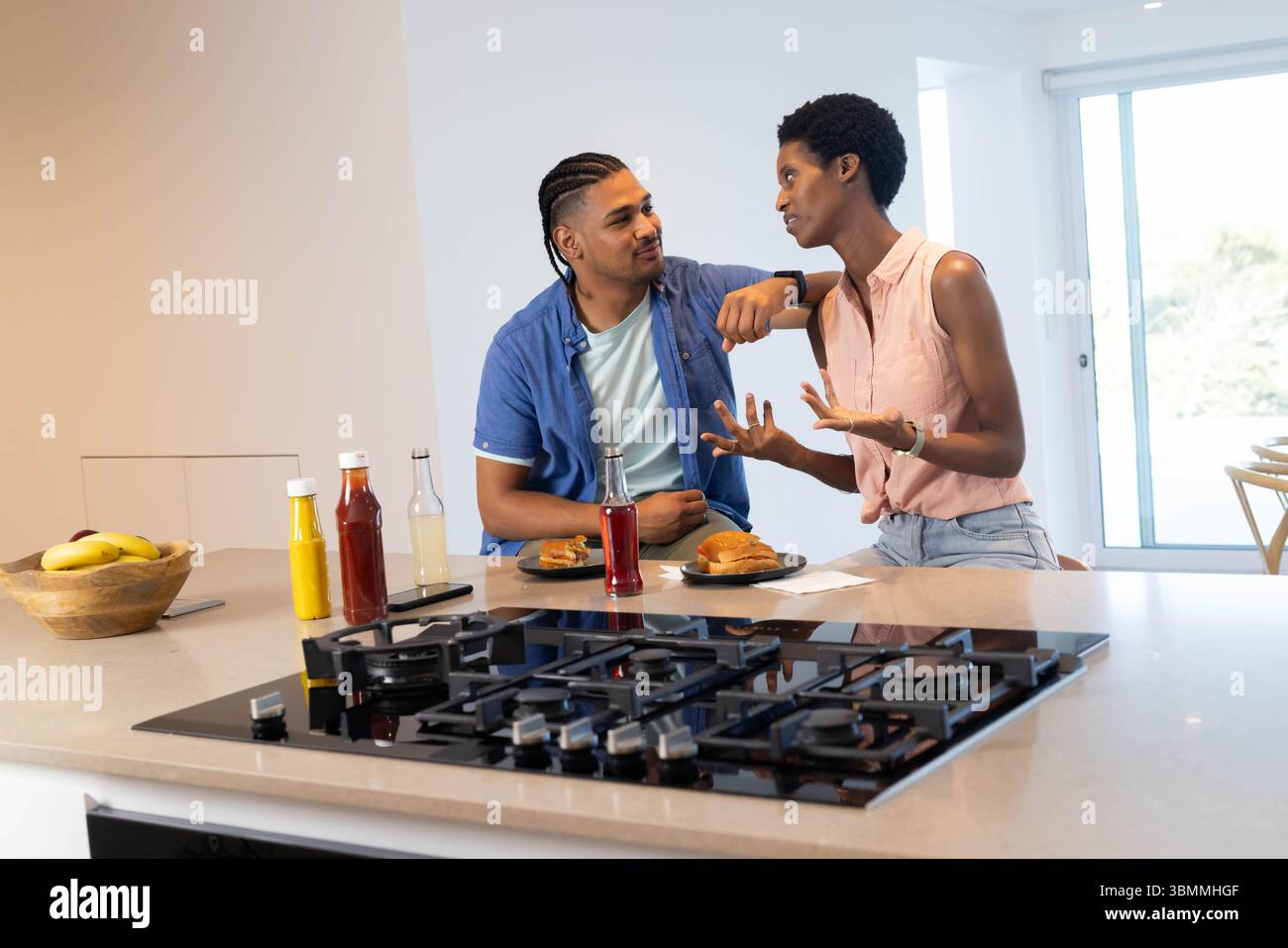 Bavardage couple diversifié s'appuyant sur le comptoir en pierre de cuisine moderne, avec plaque de cuisson à gaz et sandwichs Banque D'Images