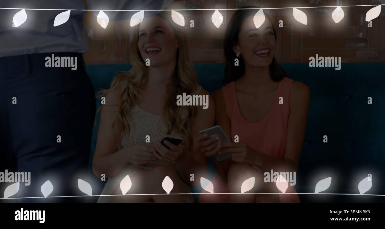 Amis assis portant de l'or, robes corail sur banc sarcelle dans le salon, avec smartphones et lumières Banque D'Images