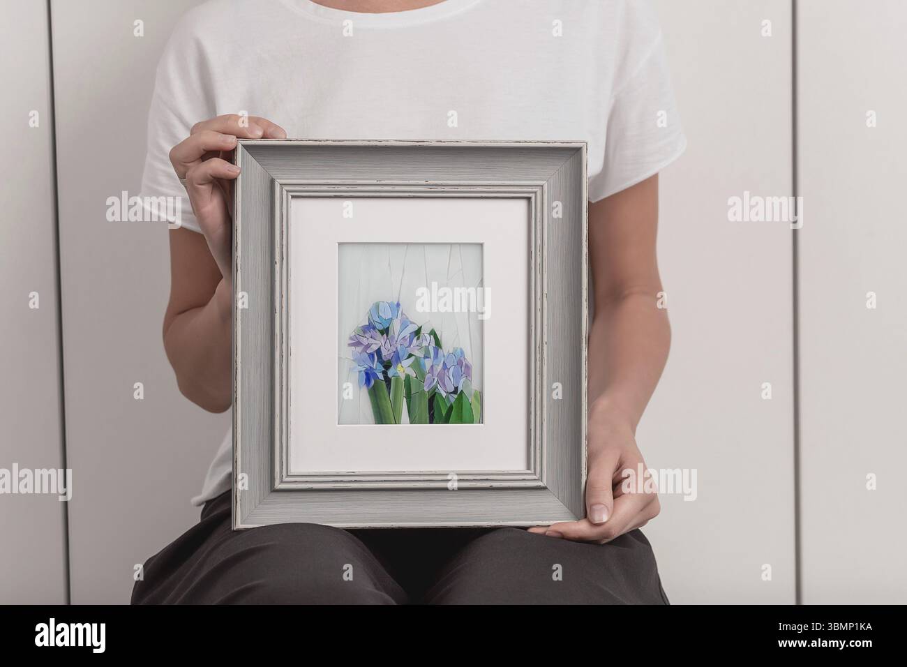 Jeune femme blonde dans un T-shirt blanc est debout dans un petit atelier et tenant en mains une mosaïque photo dans un cadre. Close-up Banque D'Images